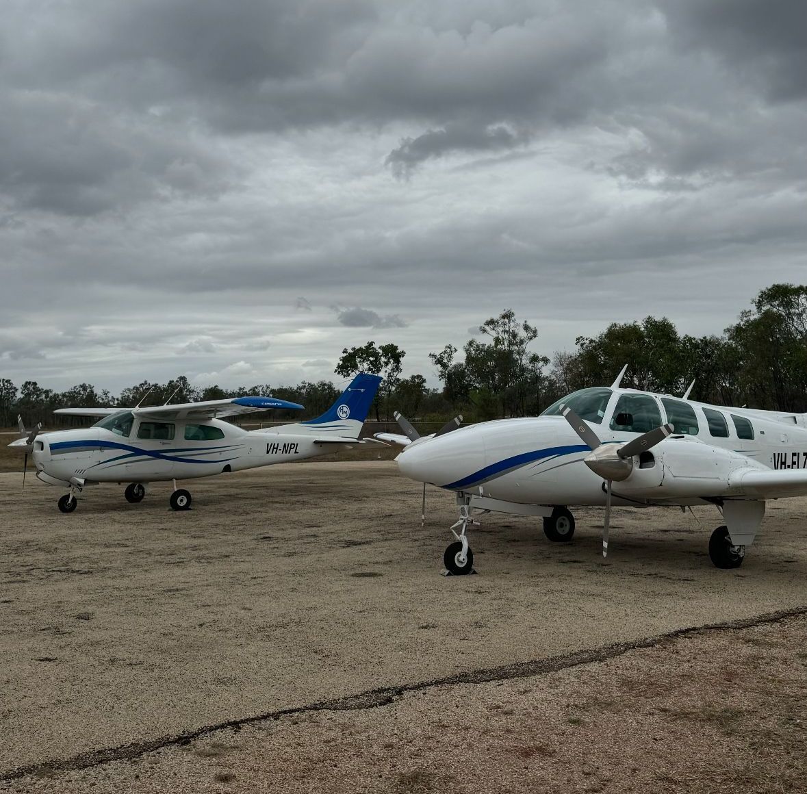 Two small airplanes on a dirt airfield under a cloudy sky. One is a twin engine plane.
