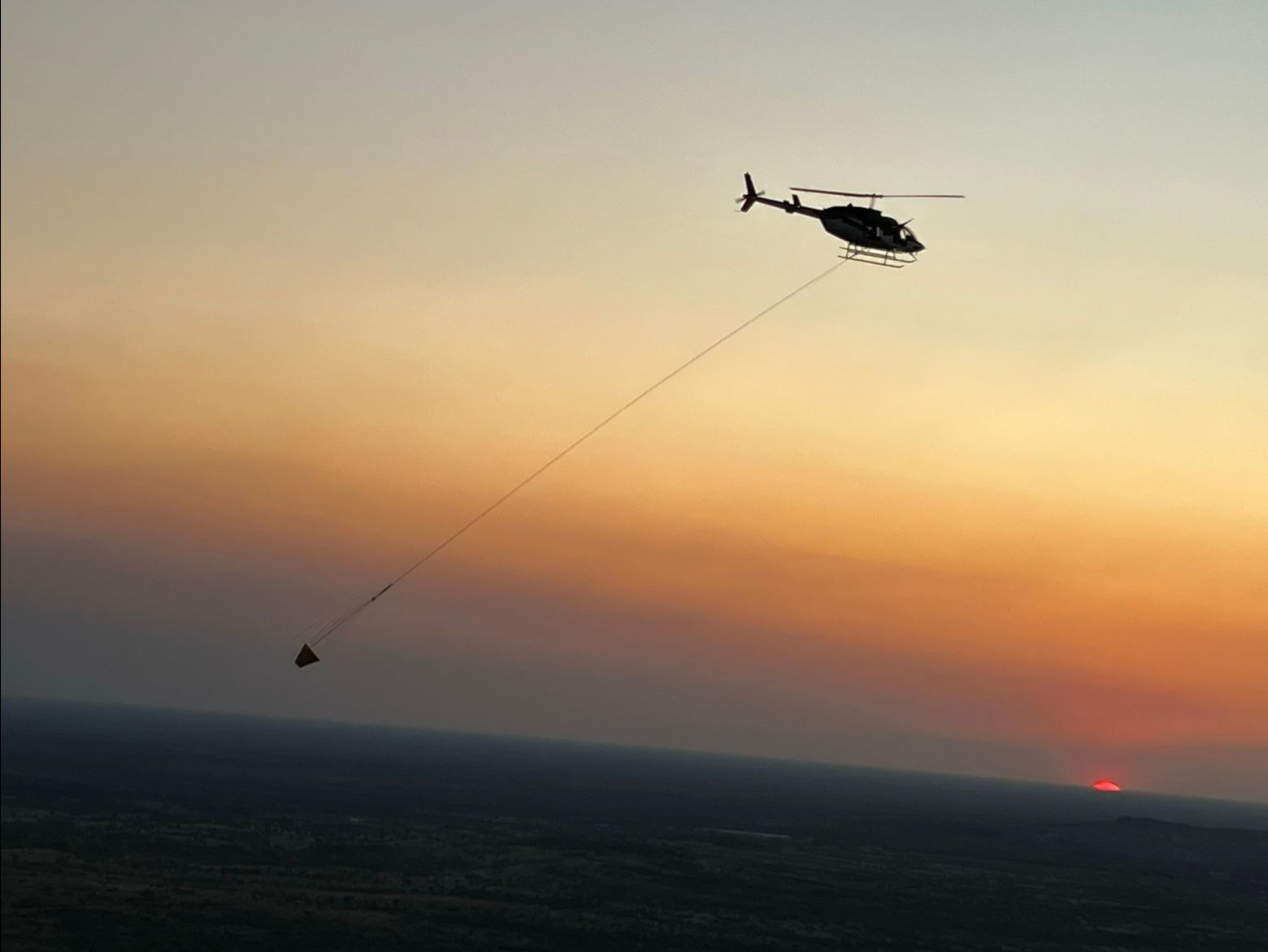 Helicopter flying with a bucket of water over a landscape at sunset; fire fighting.