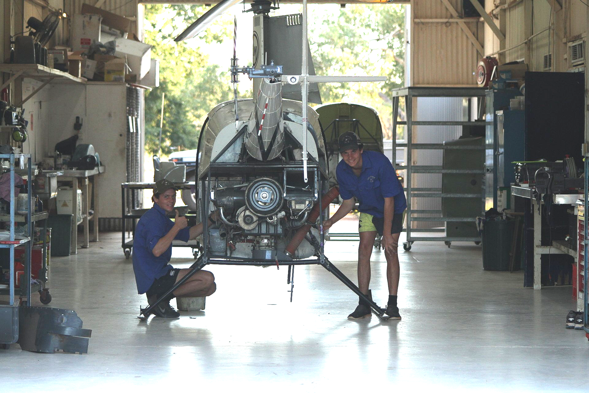 Two people working on a helicopter inside a hangar. One kneels, while the other stands, both wearing blue.
