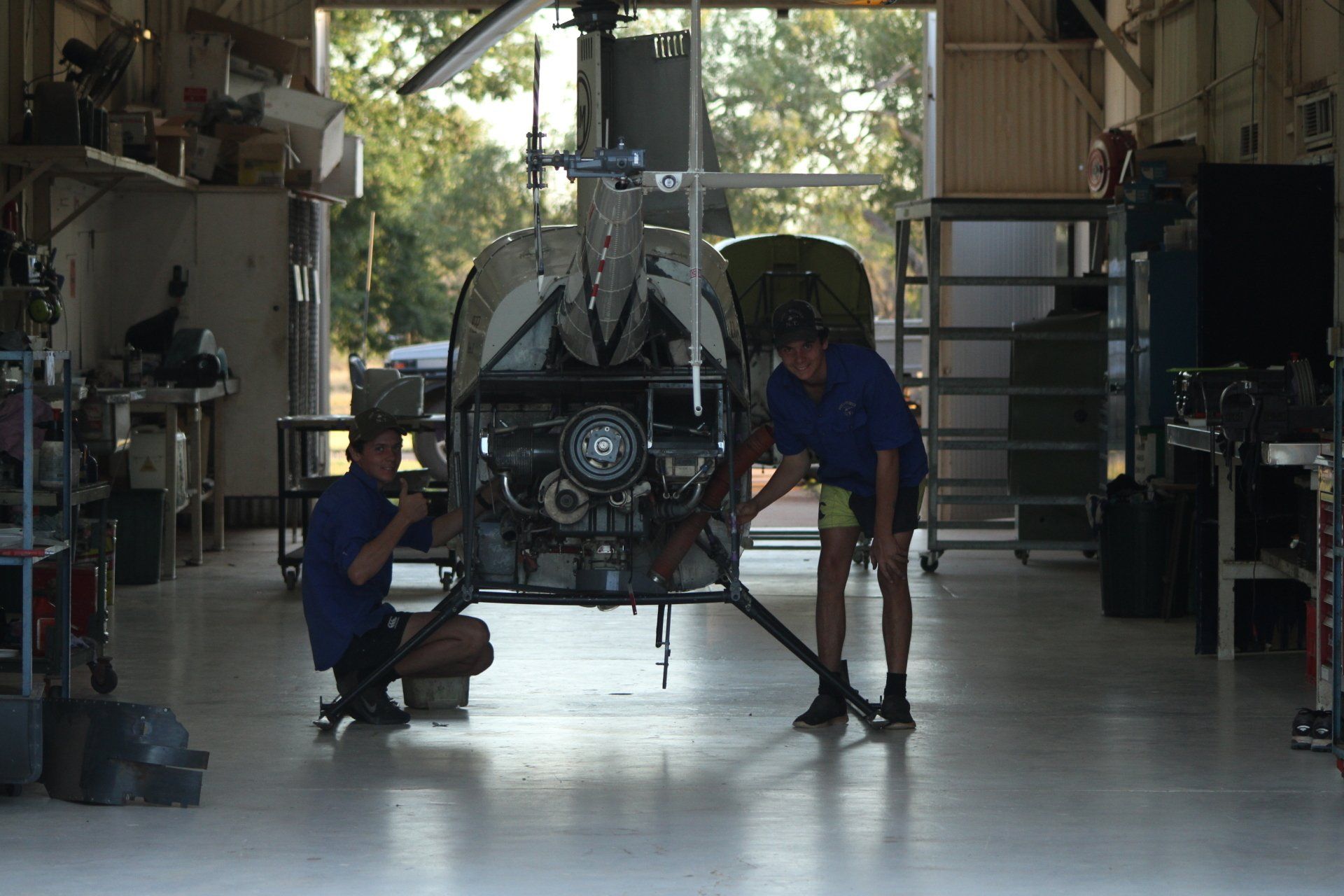 Two people working on a helicopter inside a garage, lit by natural light.