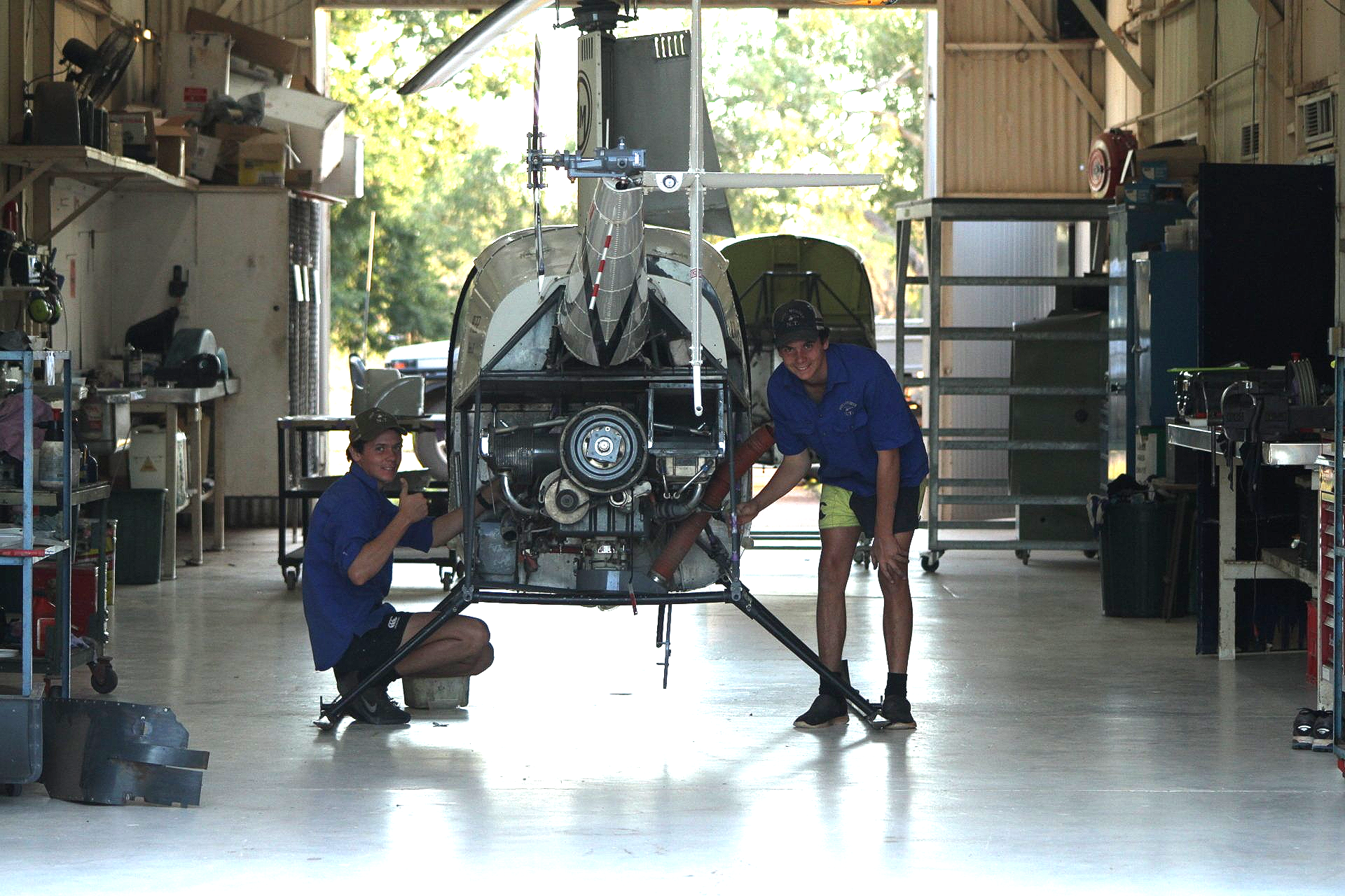 Two people working on helicopter in a hangar. One kneeling, one standing; both wearing blue shirts.