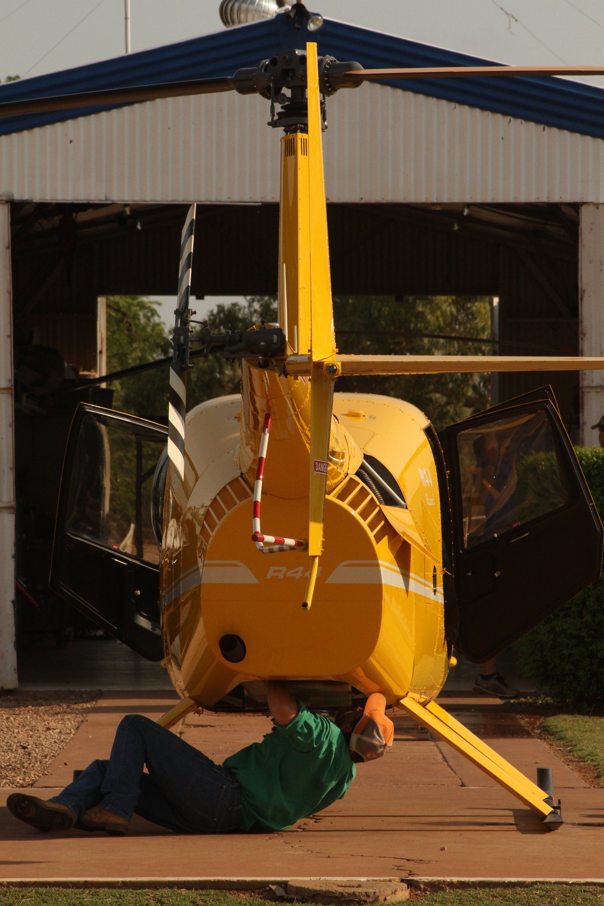 Person lying under a yellow helicopter for maintenance, in front of a hangar.