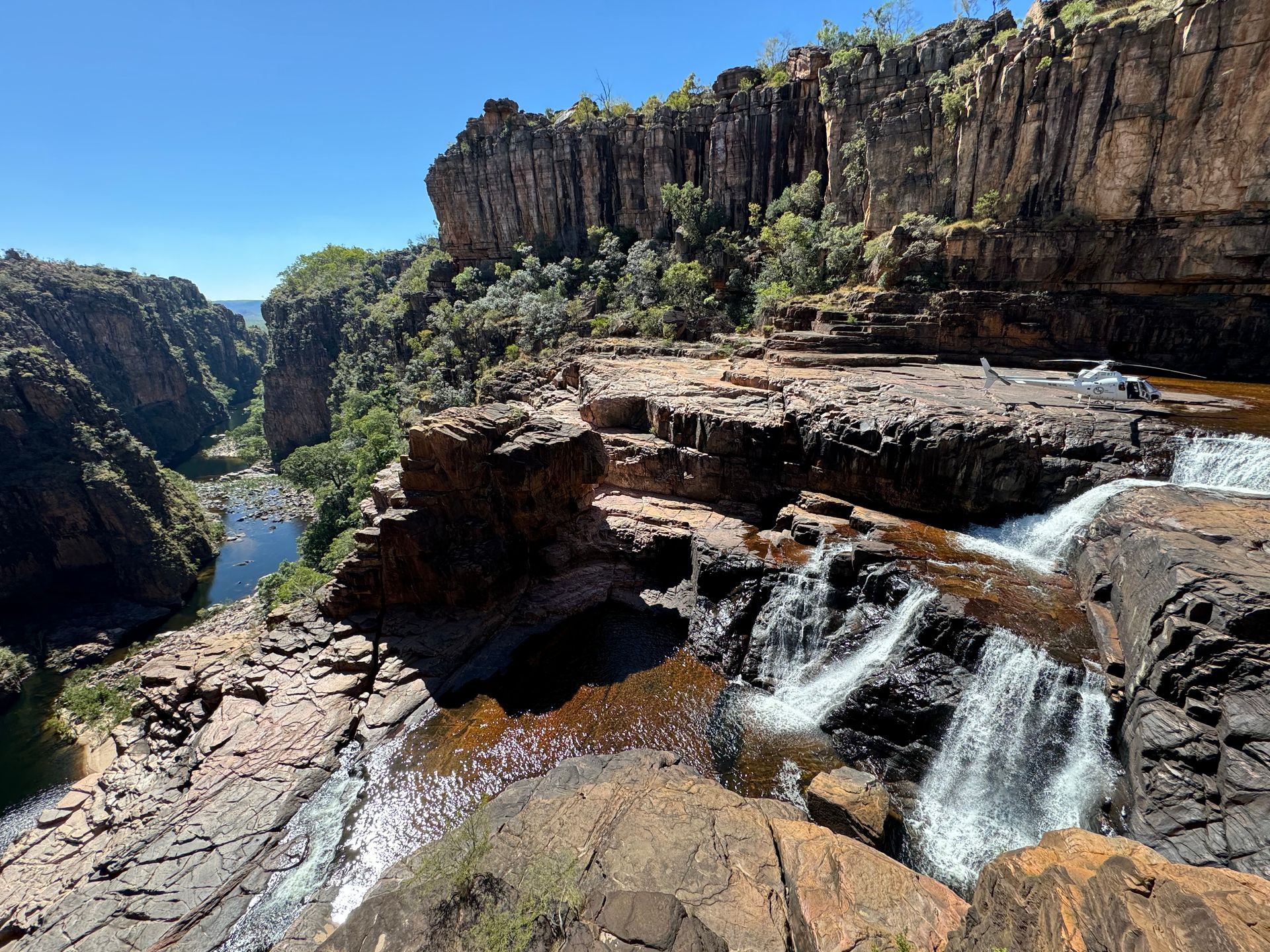 Waterfall cascading over rocky terrain into a river, with high canyon walls under a blue sky.