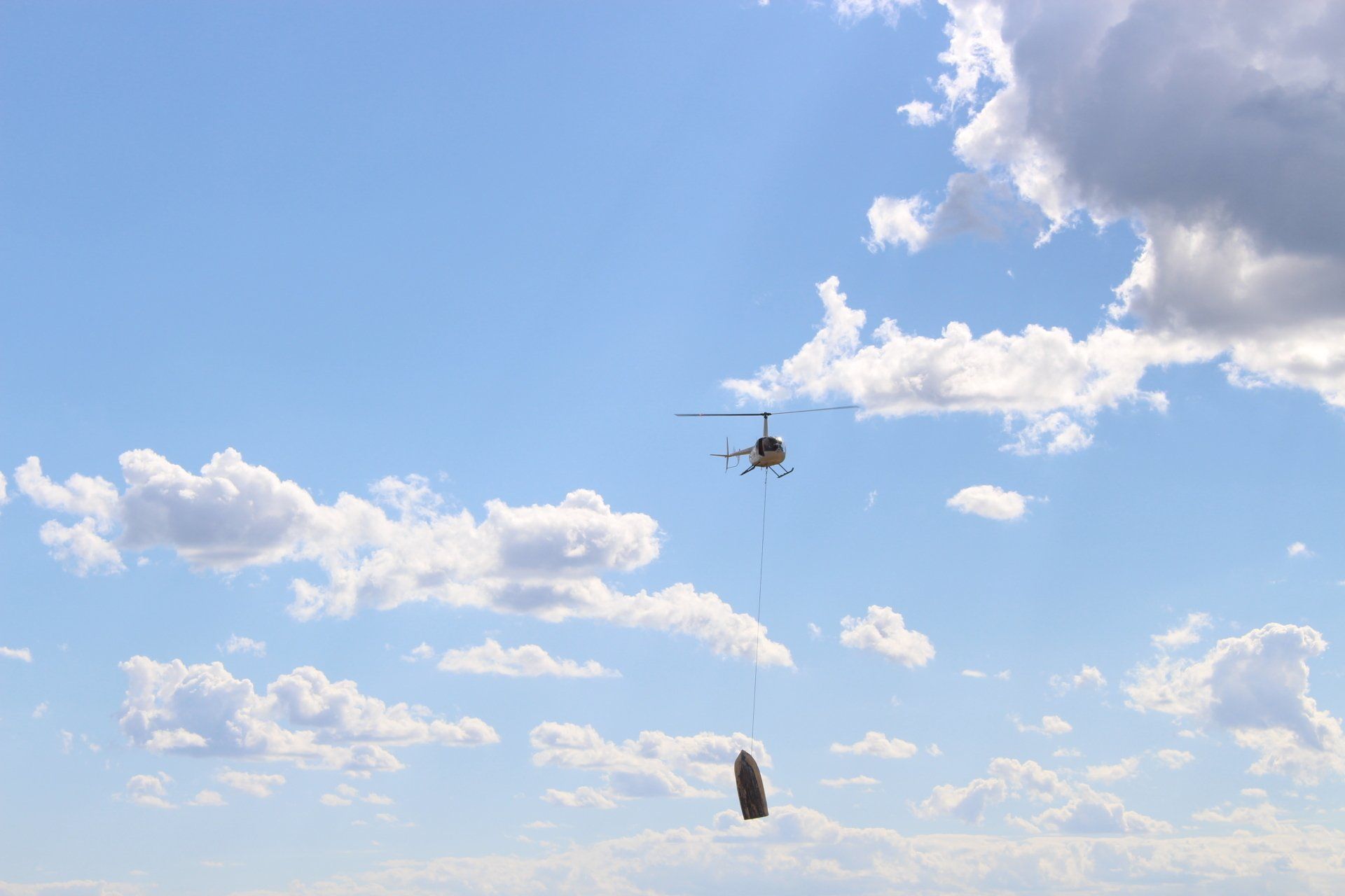 Helicopter flying in a blue sky with clouds, carrying a brown object below.