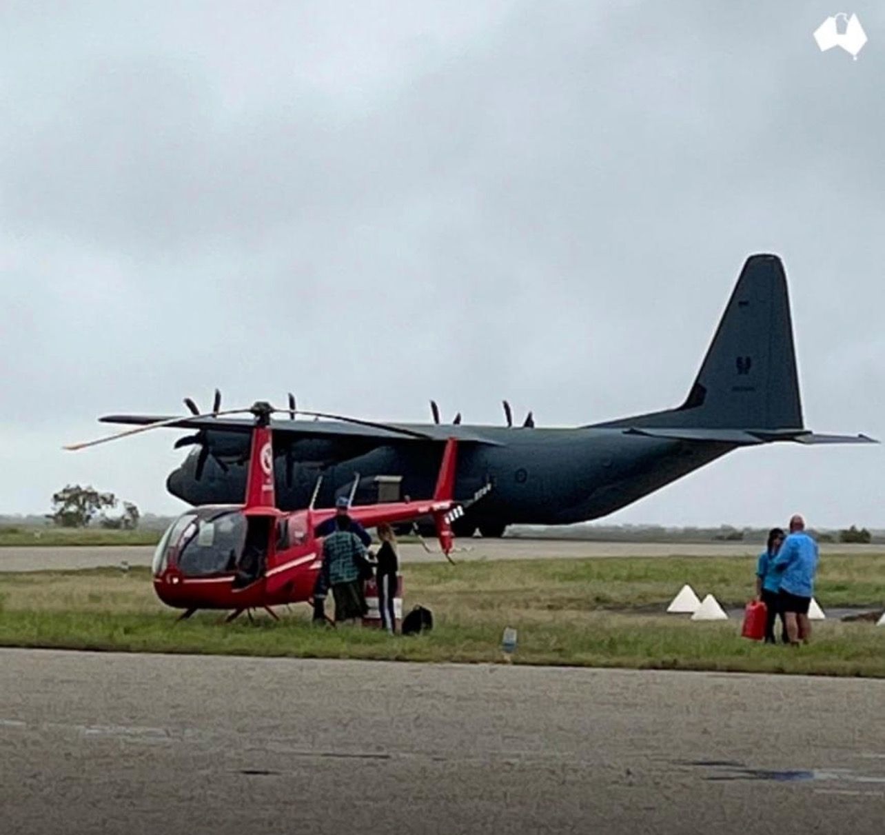 Red helicopter parked next to a large dark gray aircraft on an airstrip with people nearby.