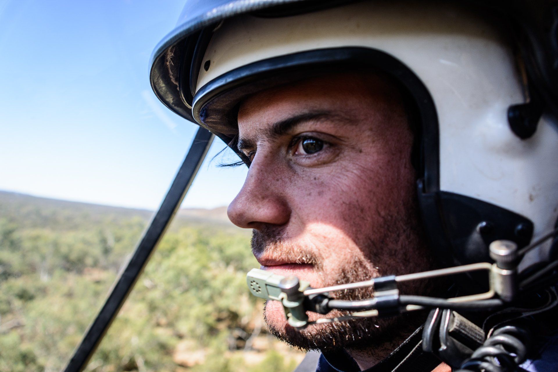 Man in a helicopter cockpit, wearing a headset and helmet, looking intently out the window.