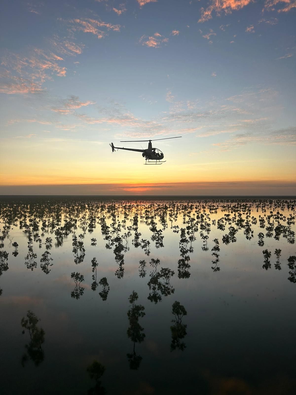 Helicopter flies over a still body of water dotted with trees during a colorful sunset.