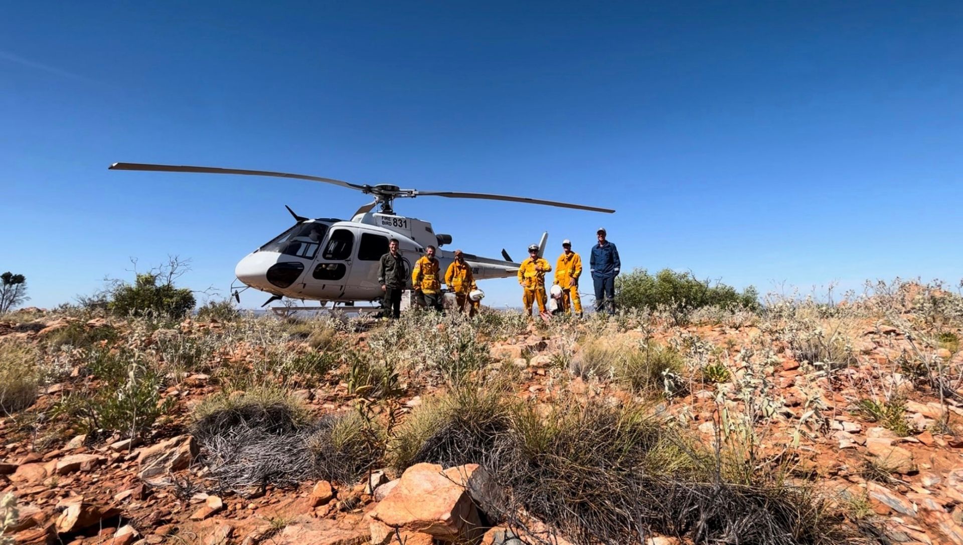 Helicopter with three people in yellow suits on a rocky, dry hillside on a sunny day.