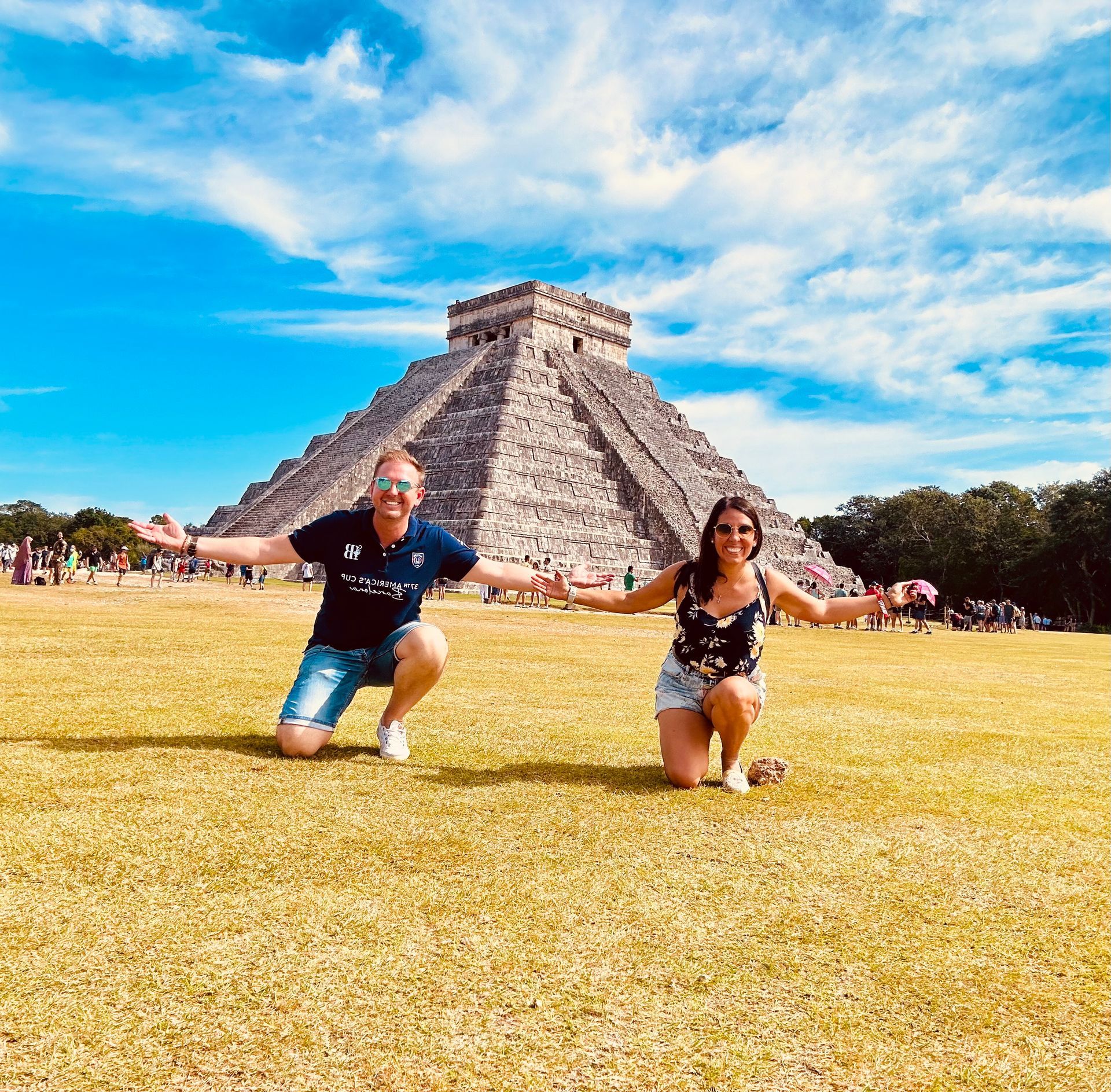 Dos personas arrodilladas con los brazos extendidos frente a la pirámide de Chichén Itzá, bajo un cielo azul.