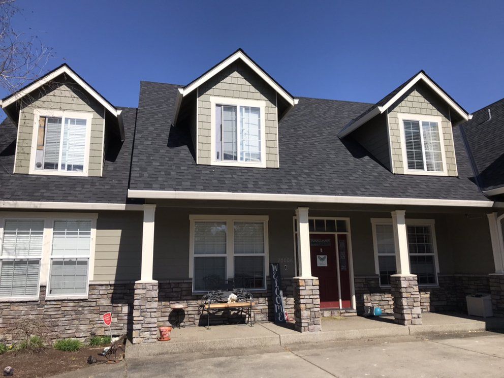 Multi-story house with dark gray roof, three dormers, tan siding, and stone accents.