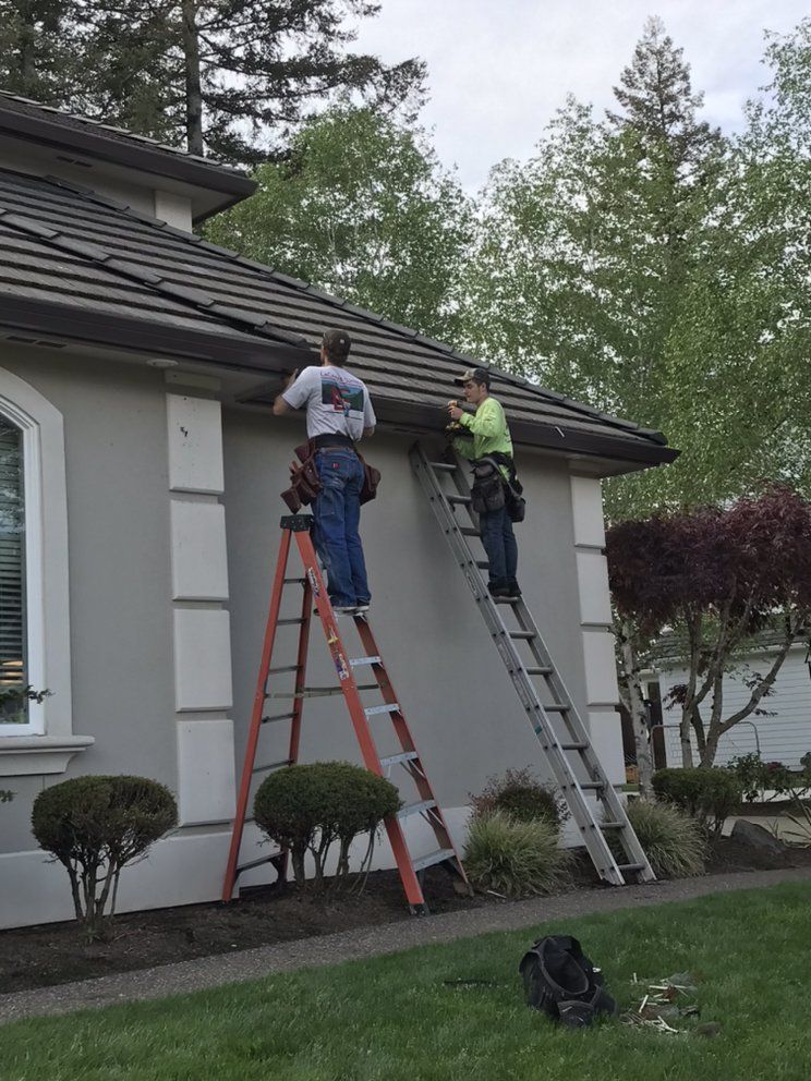 Two people on ladders fixing a roof's gutter on a light-colored house. Green grass and trees are in the background.