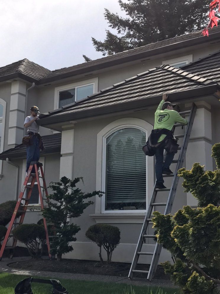 Two workers on ladders cleaning gutters of a house. One reaches up, other on lower ladder.