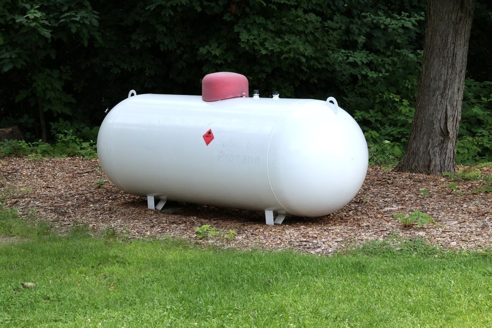 White propane tank on a gravel bed next to grass and trees.
