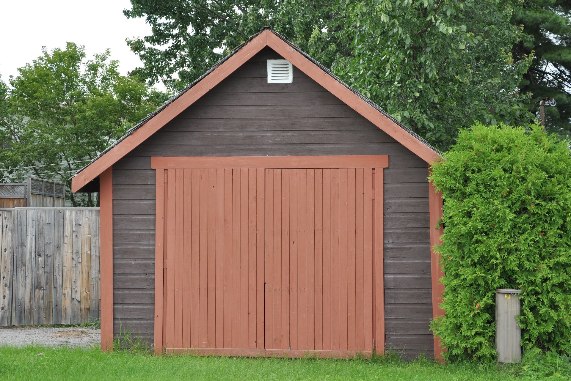 Small red wooden shed with double doors in a grassy yard, backed by trees and a fence.