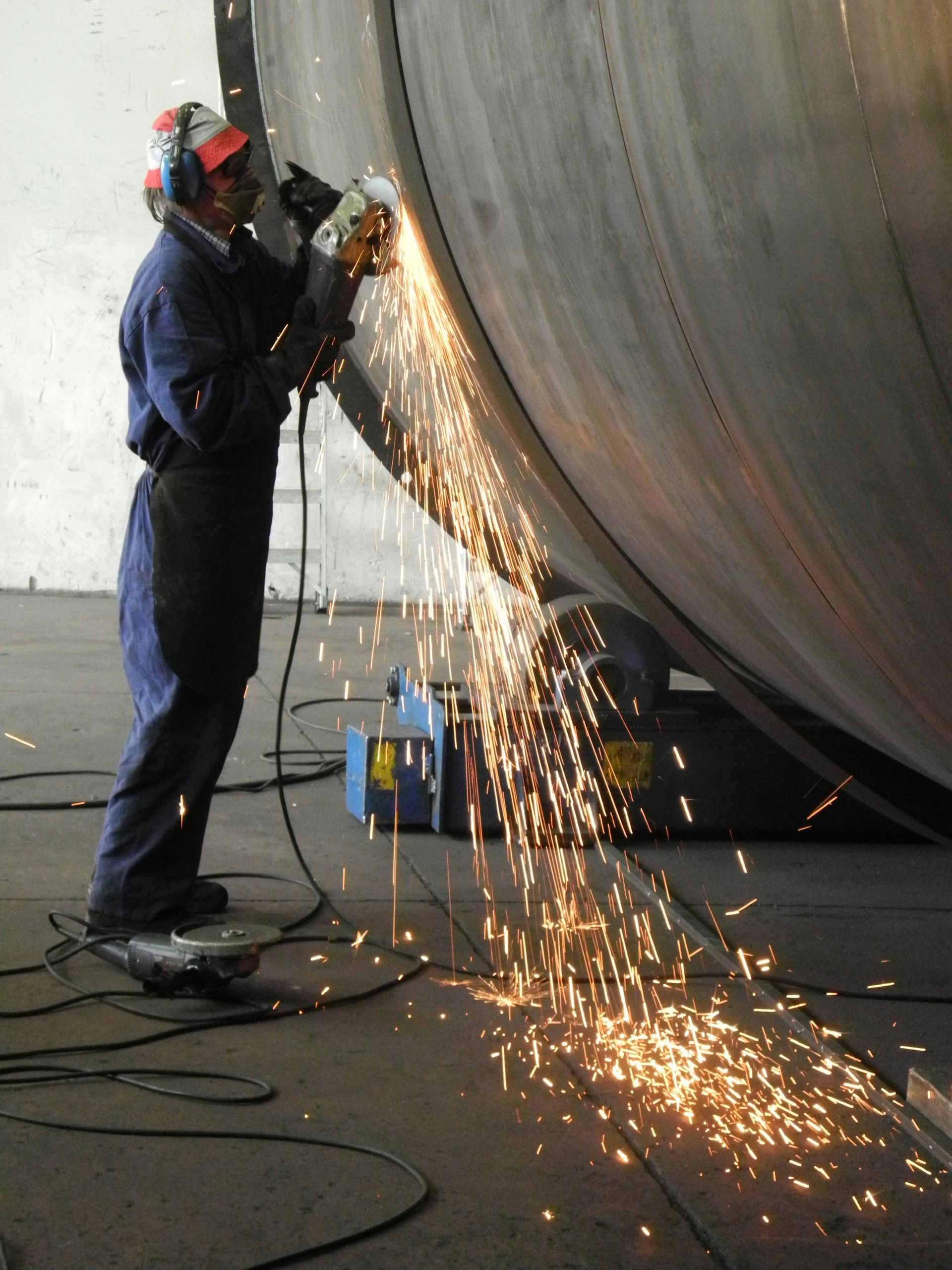 Person in protective gear grinding a large metal cylinder, sending bright sparks across the floor.
