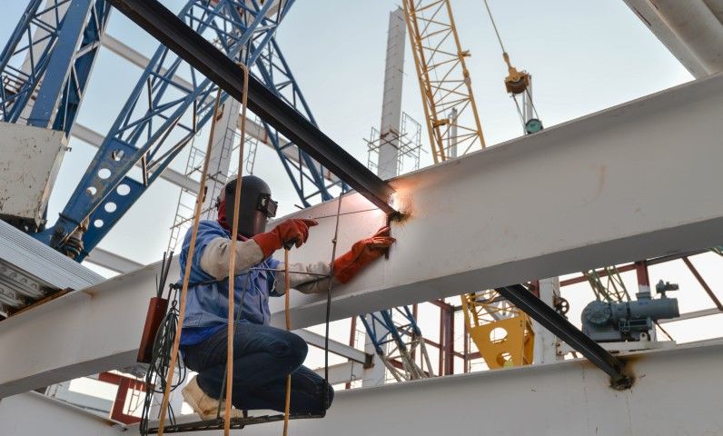 Construction worker welds steel beam, wearing protective gear, with a crane in the background.
