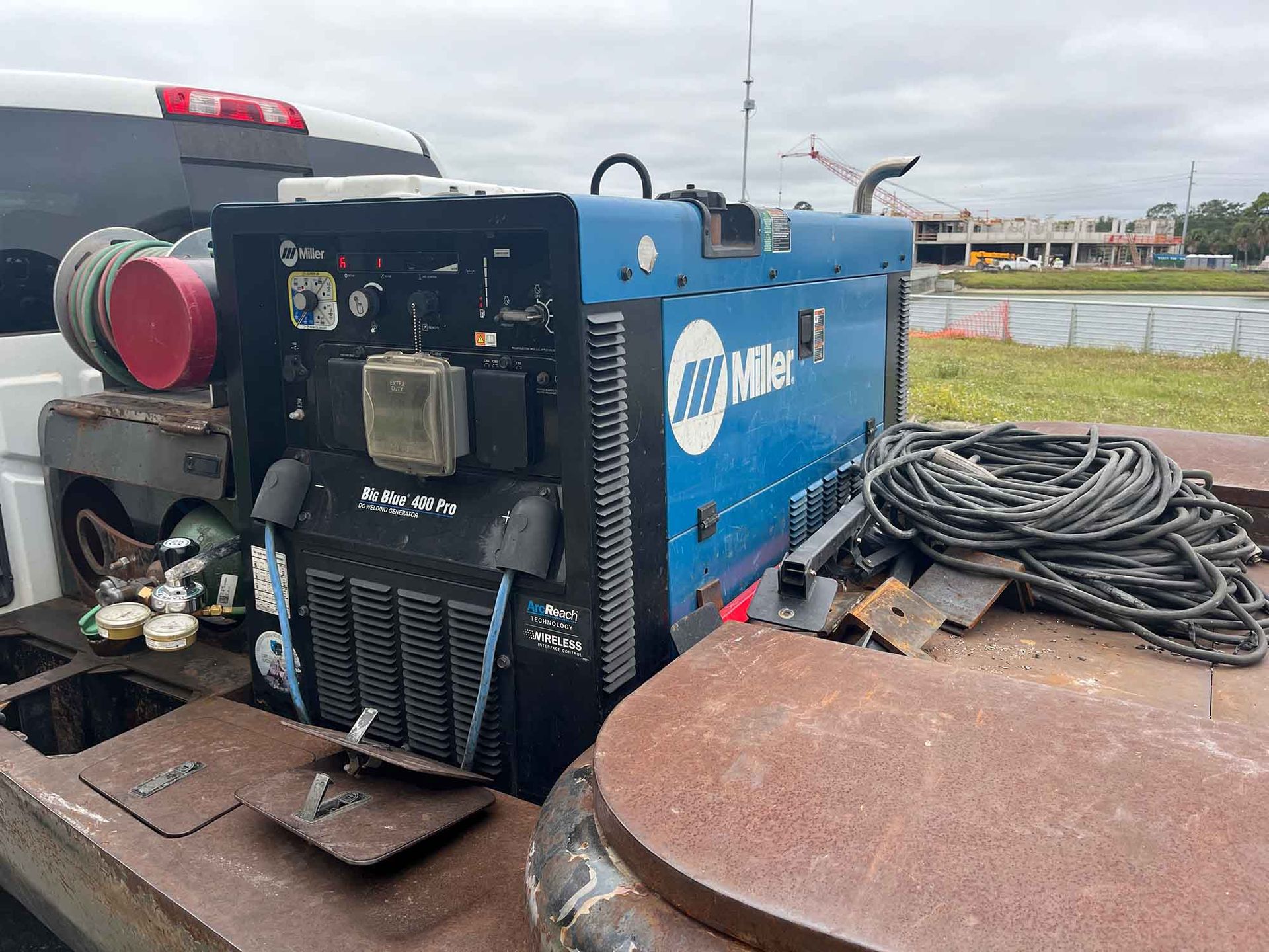 Blue Miller welding machine on a rusty truck bed with coiled cables and a red tank in an outdoor worksite