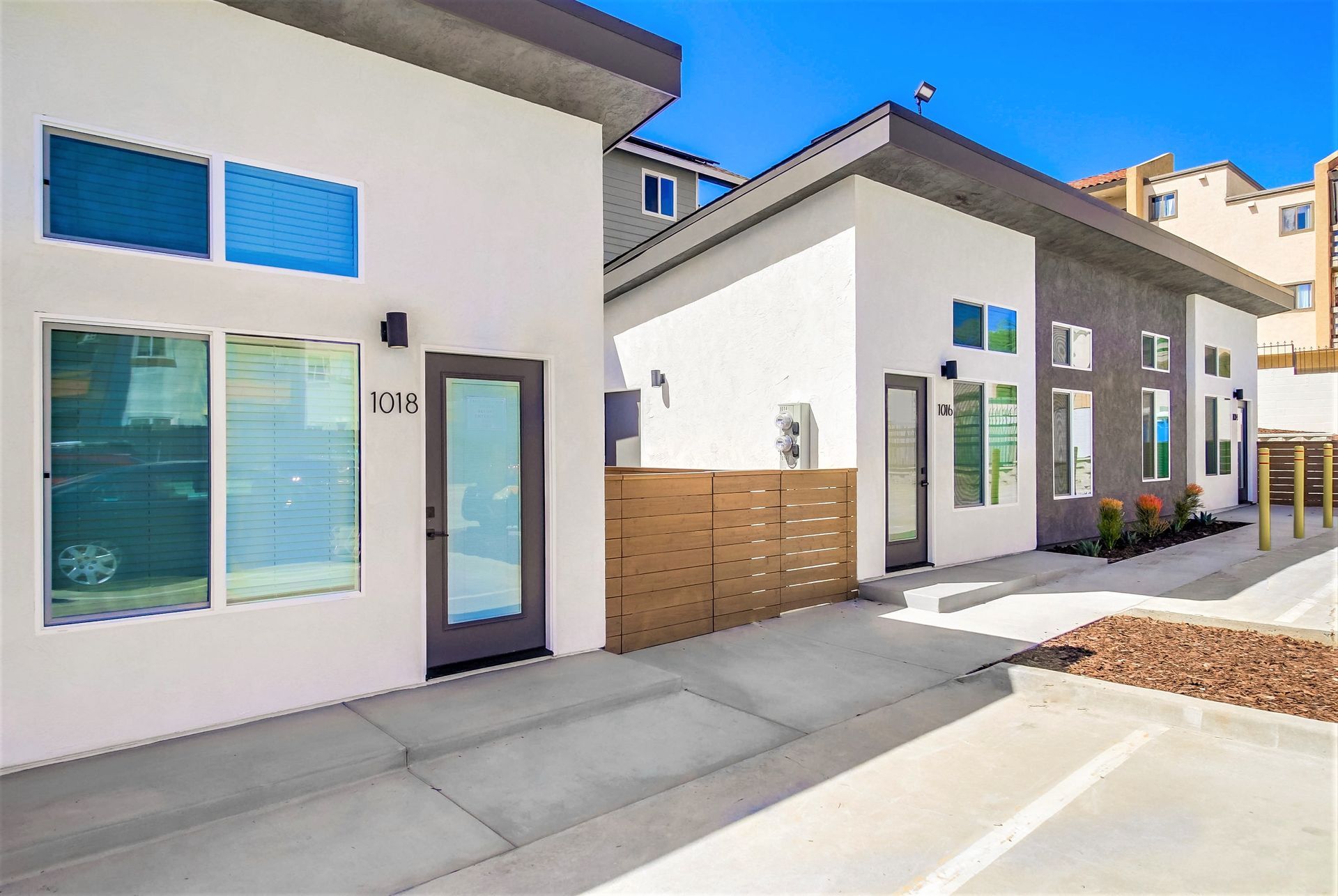 Modern townhouses with cream-colored siding, dark gray accents, glass-panel doors, and concrete walkways under a blue sky.