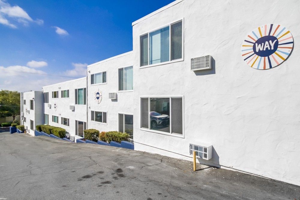 A multi-story white apartment building under a clear blue sky, featuring a colorful circular logo labeled