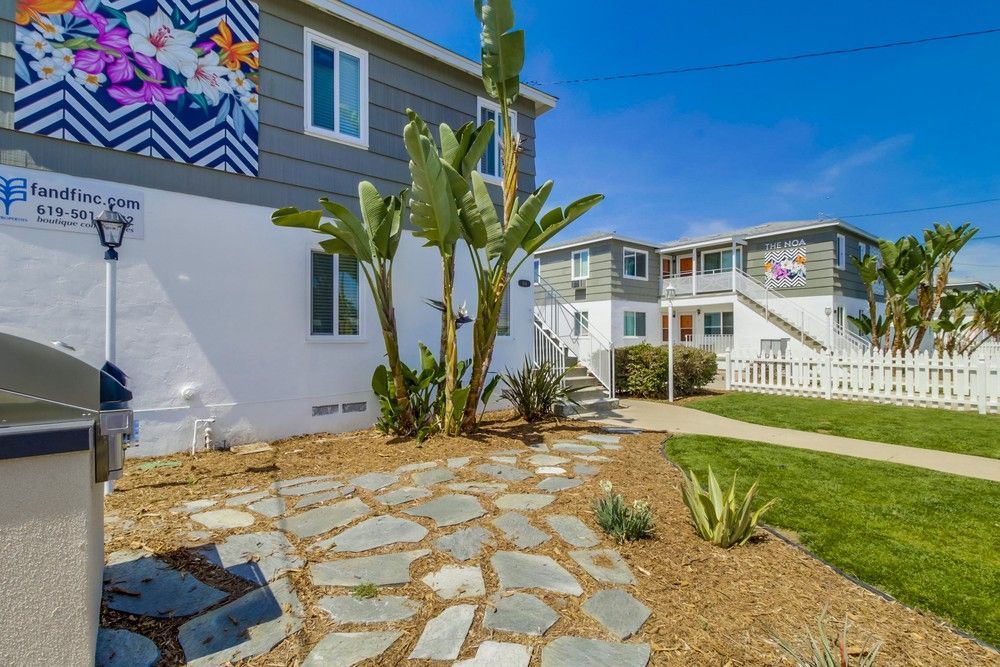A low-angle view of two-story apartment buildings with white stucco, gray trim, palm plants, and a flagstone pathway.