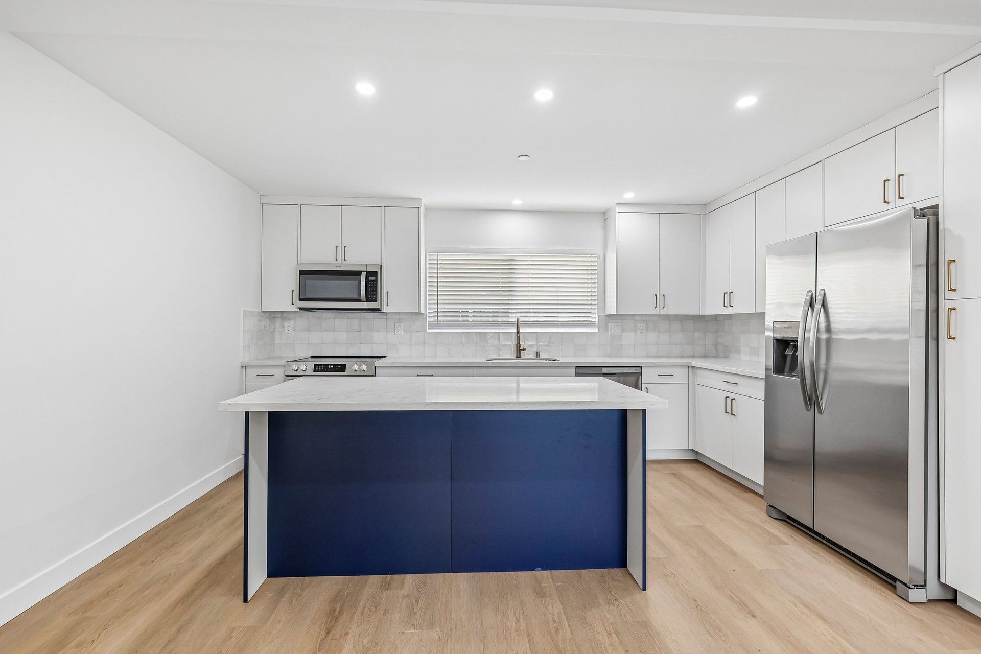 A modern kitchen with a dark blue island, two pendant lights, white and brown cabinetry, and light-colored countertops.
