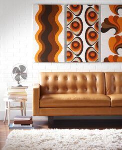 A tan leather sofa sits against a white brick wall decorated with three panels of retro, orange-and-brown patterned art.