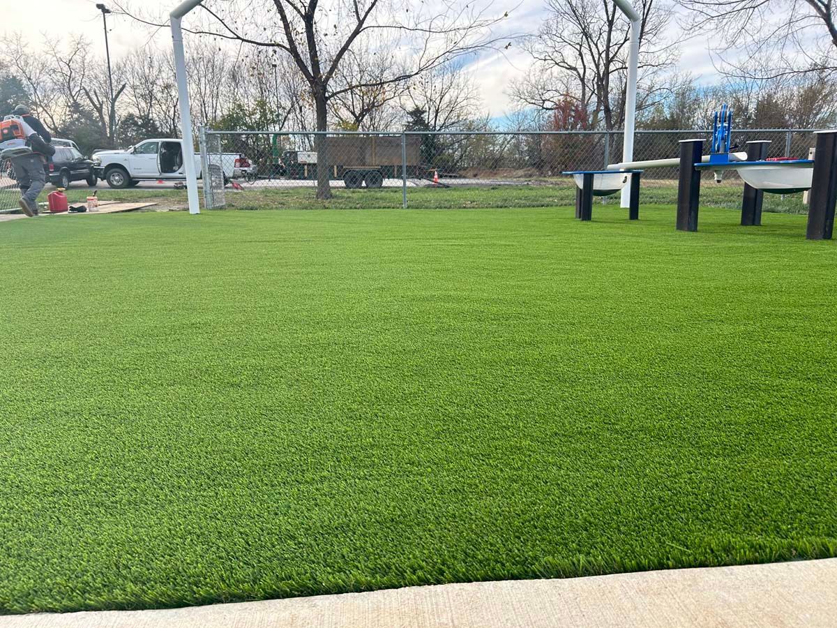 A green artificial turf playground with a fence and benches in the background.
