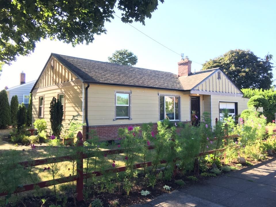 Cottage-style house with tan siding and a brick base, surrounded by a picket fence and garden.