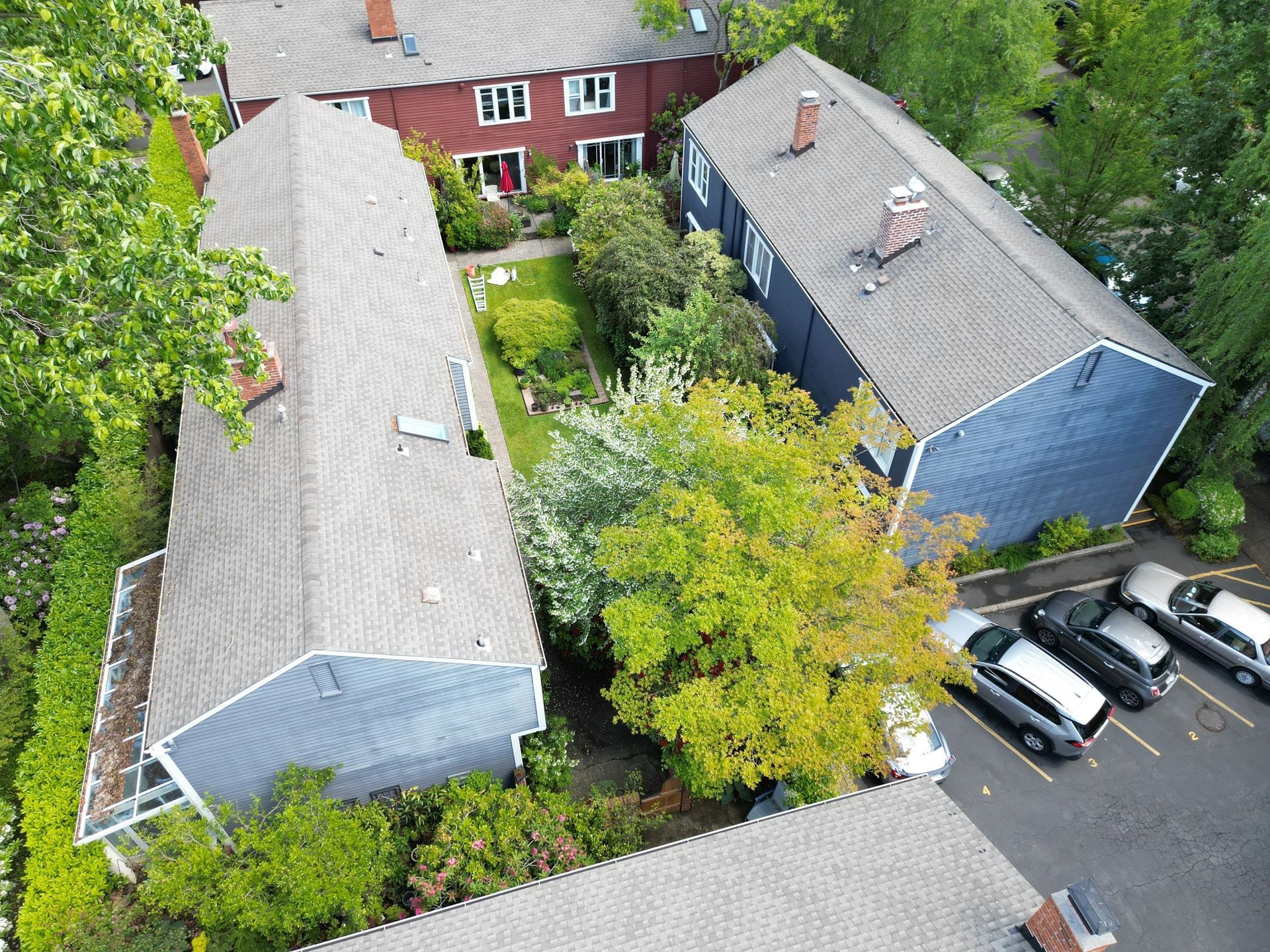 Aerial view of apartment buildings with gray roofs, trees, a small garden, and parked cars.