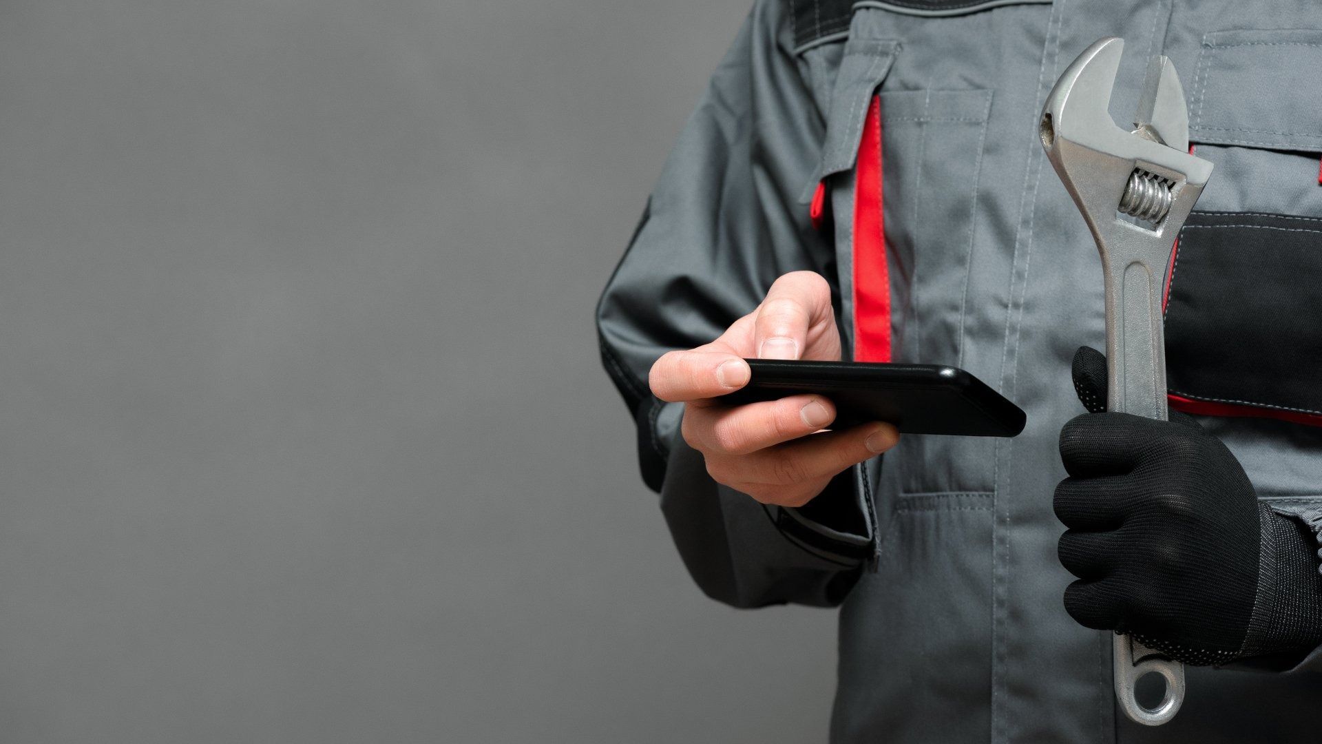 Mechanic in Gray Jumpsuit Holding a Wrench and Looking at A Smartphone Against a Gray Backdrop — Weav Plumbed in Emerald, QLD