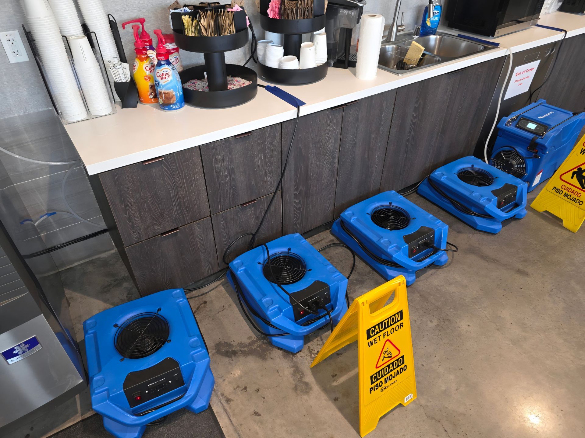 Blue water damage restoration equipment near a counter with coffee supplies and caution signs.