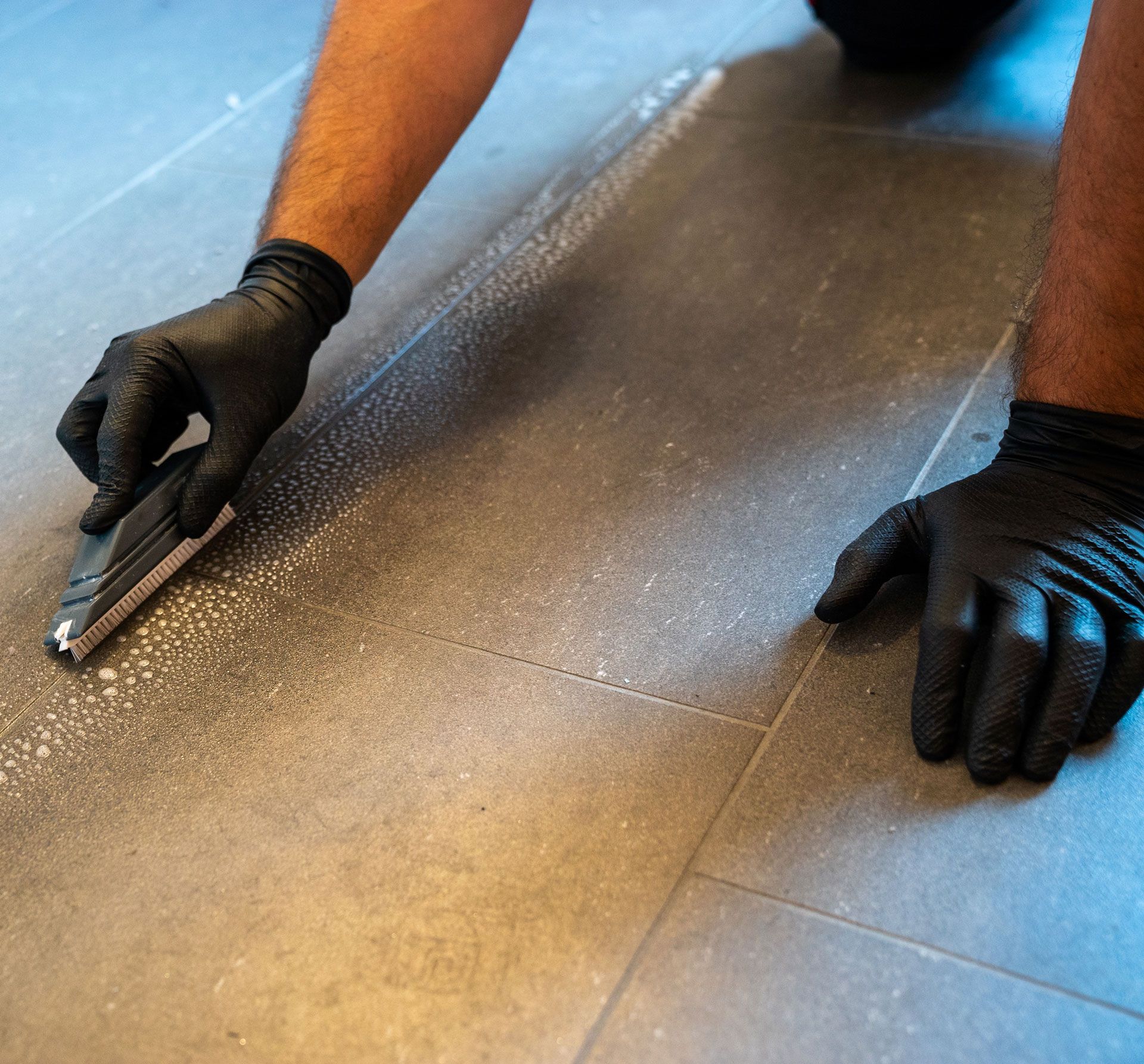 Person wearing black gloves applying grout to gray floor tiles with a trowel.
