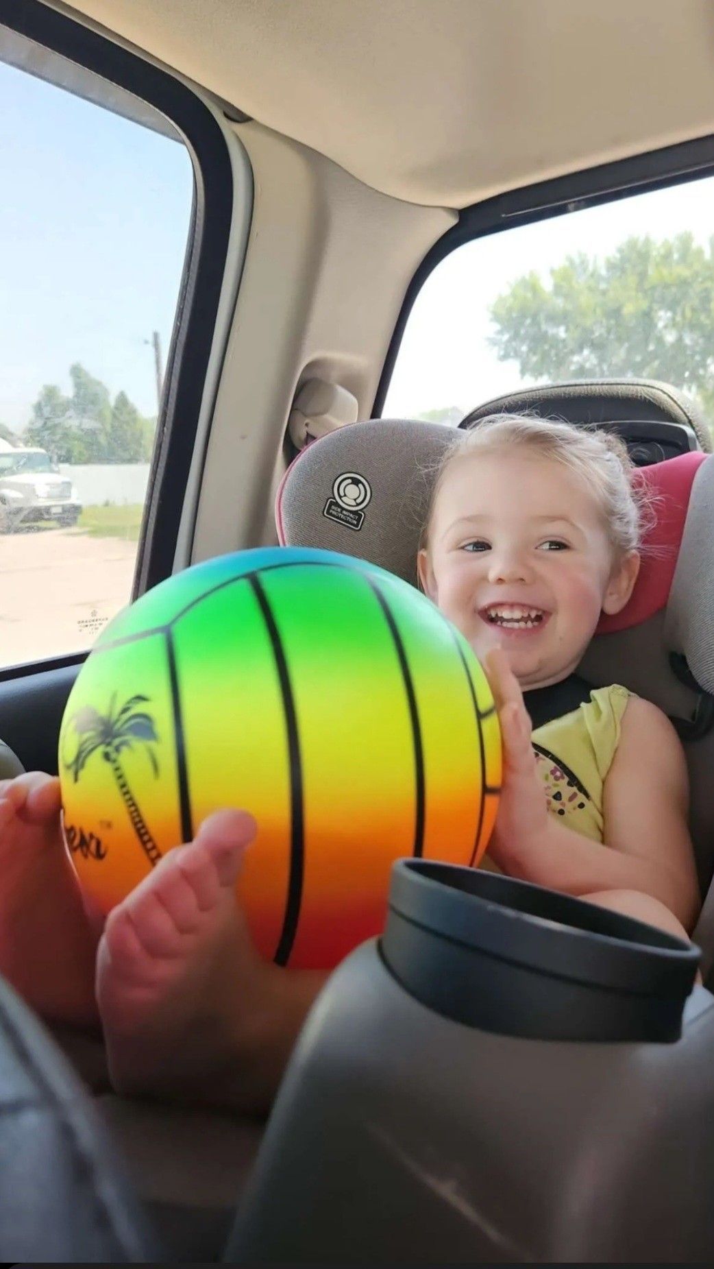 Happy toddler in car seat holding a colorful beach ball, smiling widely.