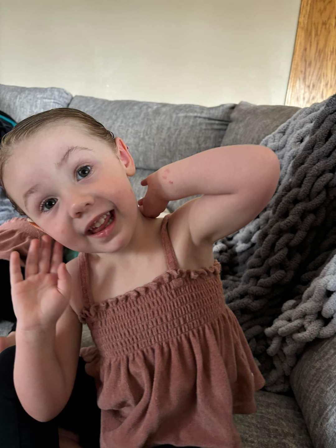 Young child with a happy expression wearing a pink top, waving, sitting on a grey couch.