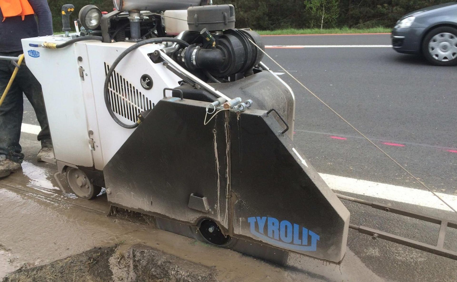 Eine Tyrolit-Betonbodensäge schneidet eine Linie in eine Asphaltstraße neben einem Arbeiter und einem vorbeifahrenden Auto.