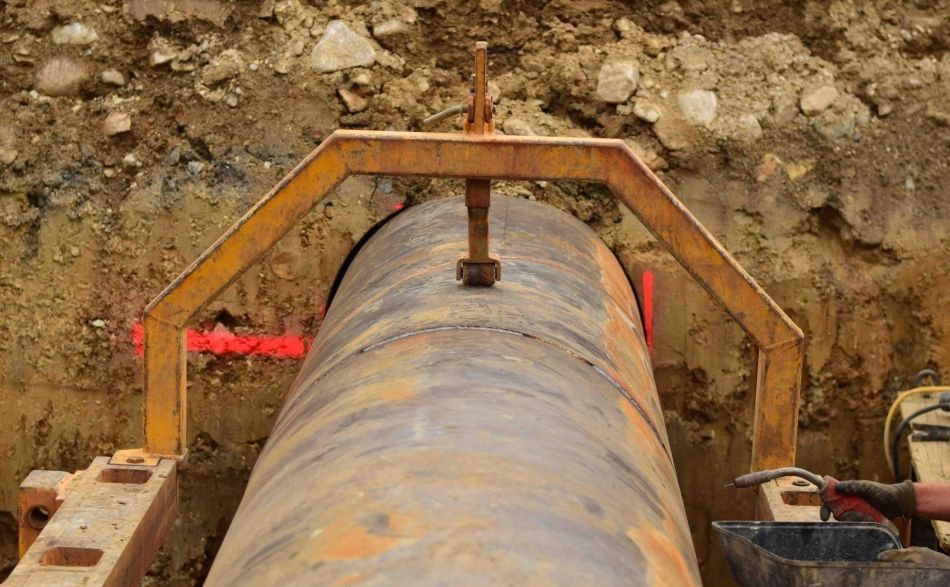 Eine gelbe Metallrohr-Ausrichtungsklemme fixiert zwei Abschnitte eines großen Industrierohrs in einem Erdgraben