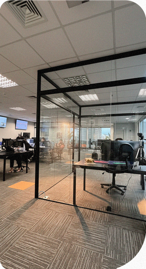 Espaço de trabalho interno com mesas, computadores e cadeiras. A luz do sol entra pelas janelas.
