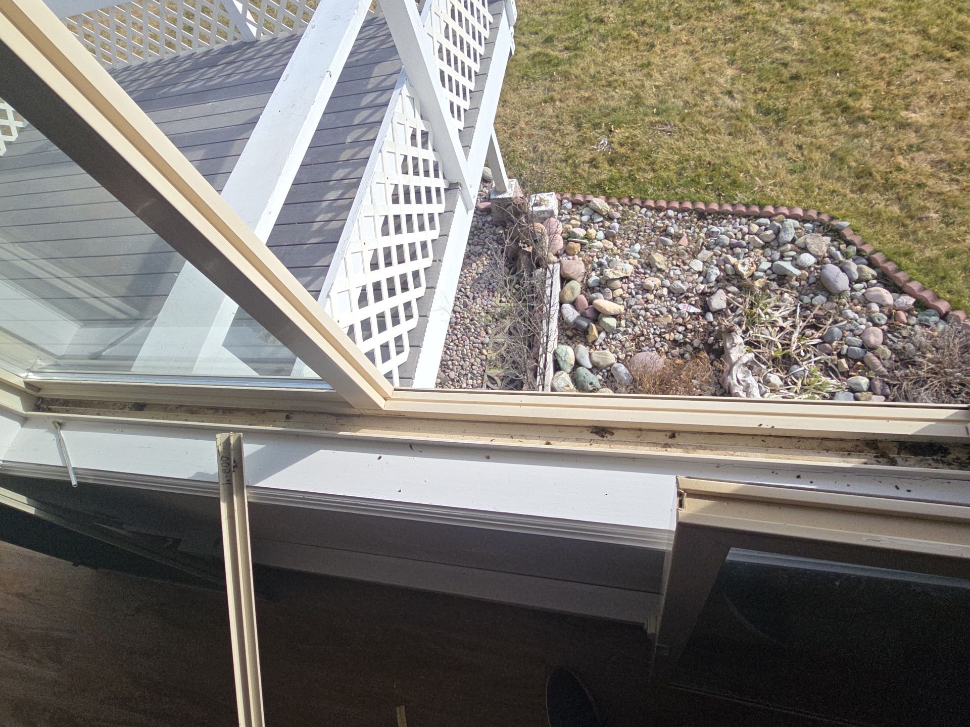 A view from inside looking out of an open window toward a white lattice porch railing and a gravel landscaping area.