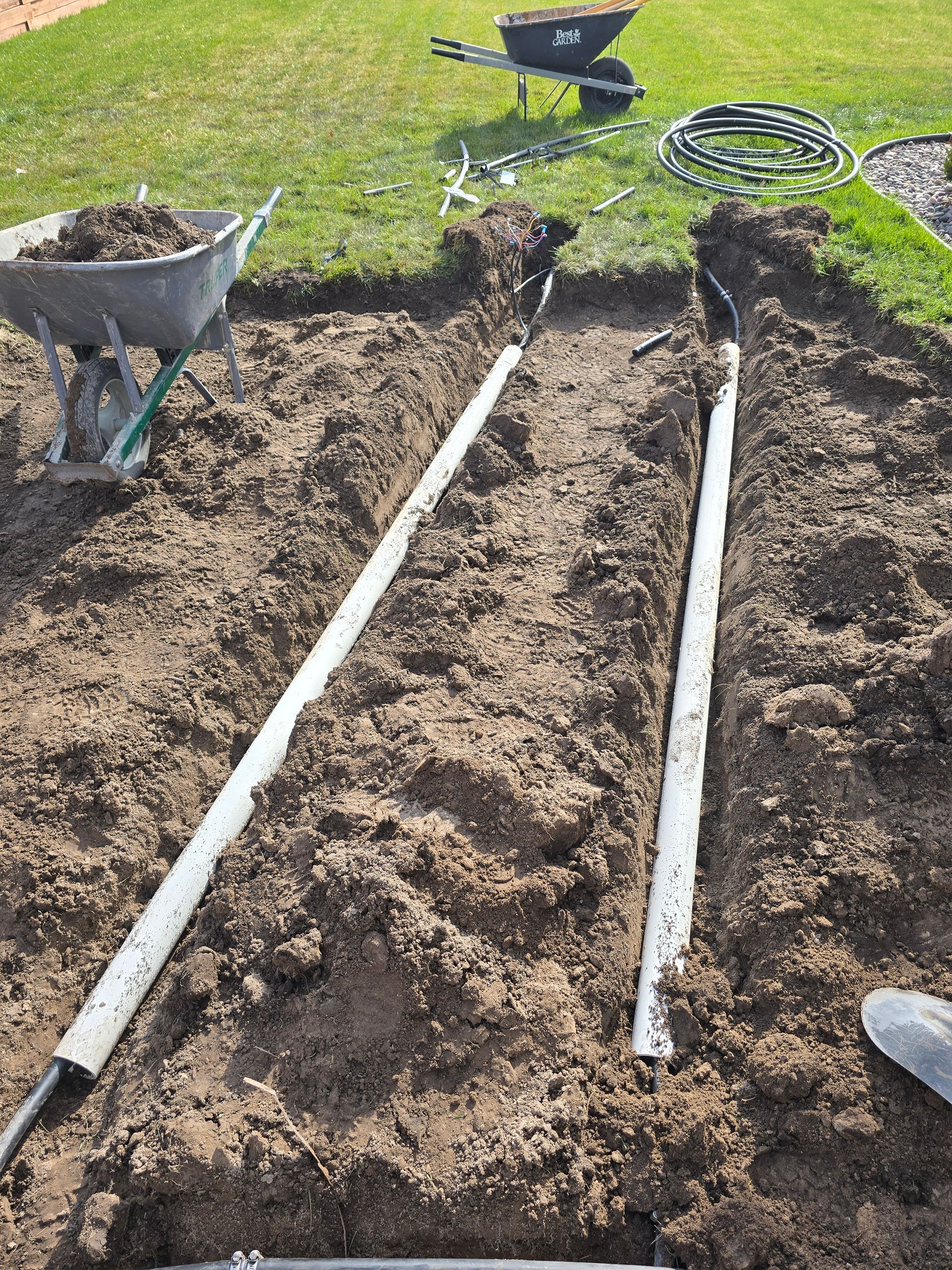Two parallel white PVC pipes are laid in dirt trenches in a yard, with wheelbarrows nearby for construction work.