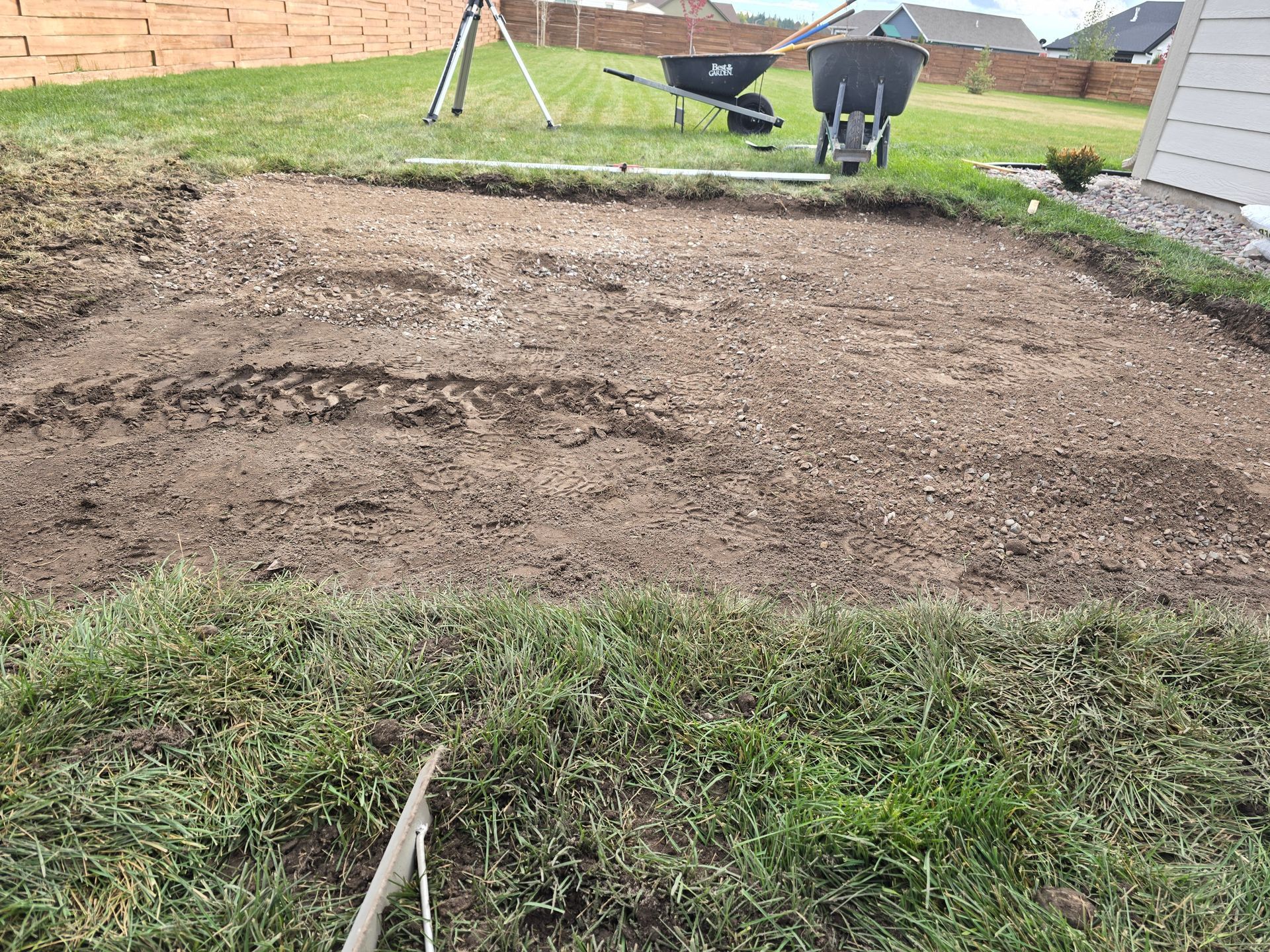 A rectangular patch of dug-up soil in a backyard, featuring a wheelbarrow and survey equipment in the background.