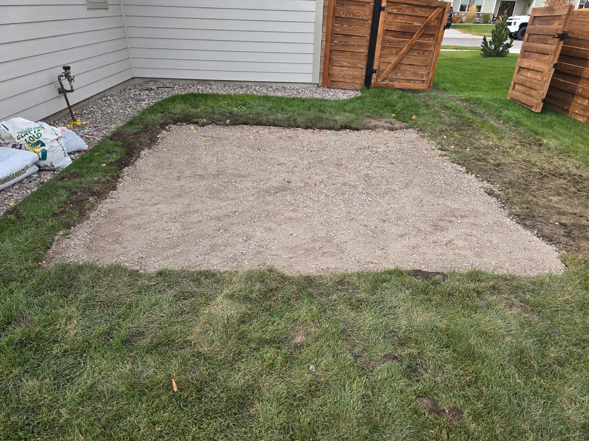 A rectangular patch of gravel framed by grass in a backyard next to a house wall and wooden fence.