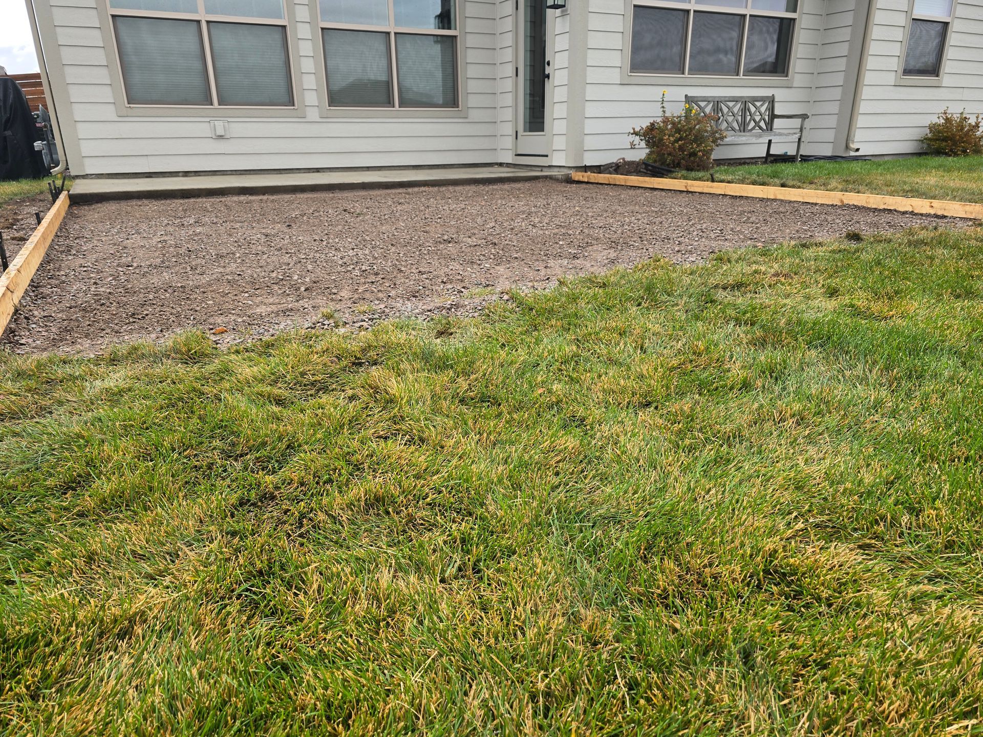 A ground-level view of a house backyard featuring a rectangular area filled with brown gravel, bordered by wooden beams.