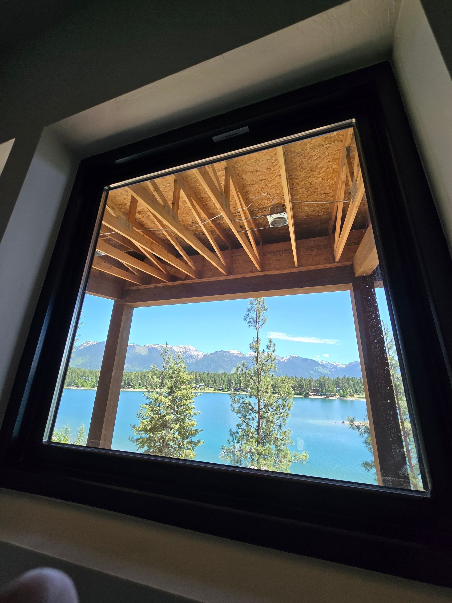A view through a square window shows a lake, trees, and snow-capped mountains beneath an exposed wooden porch ceiling.