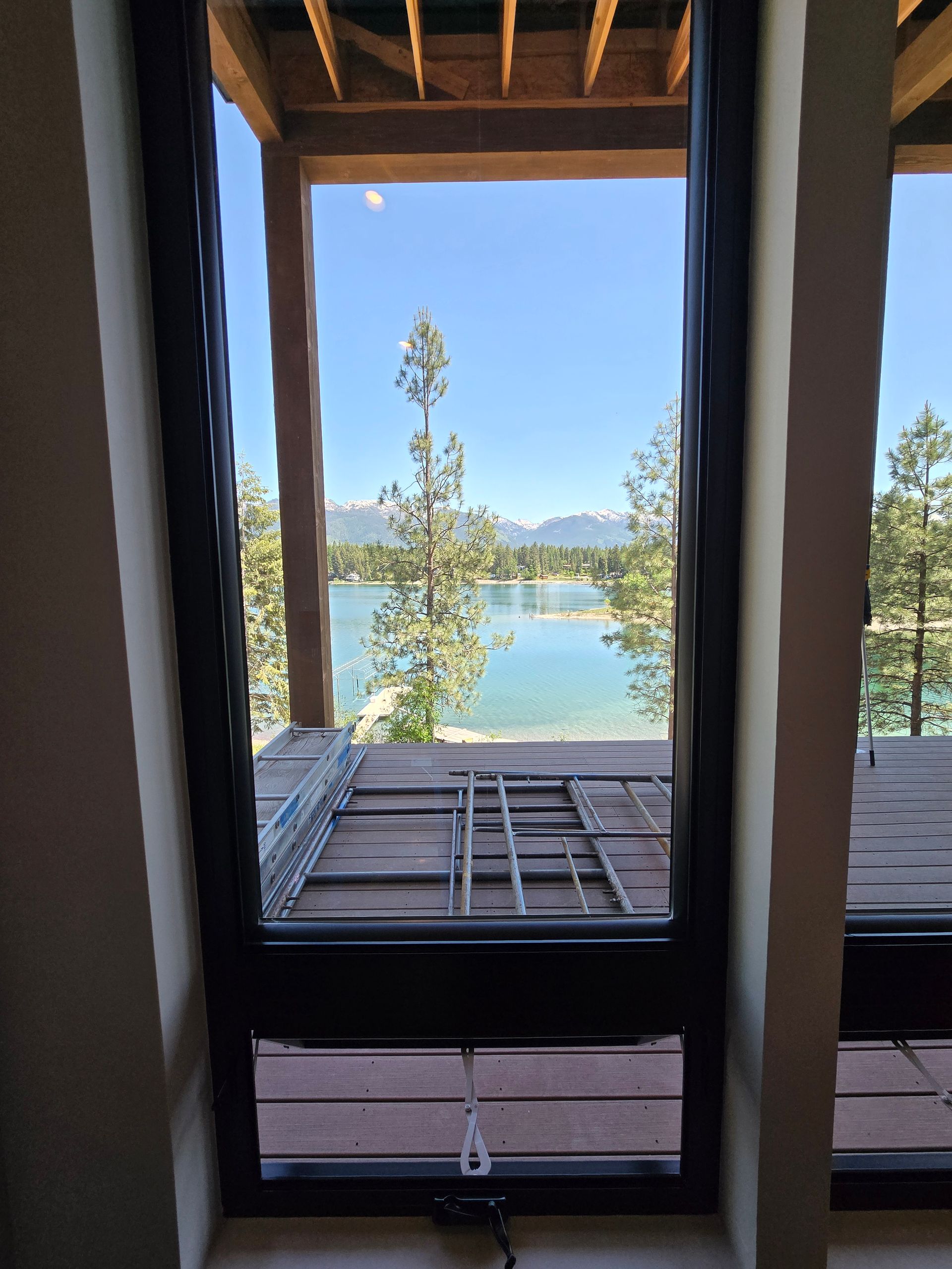 A view of a lake and pine trees framed by a tall, modern black-framed window and a wooden deck.