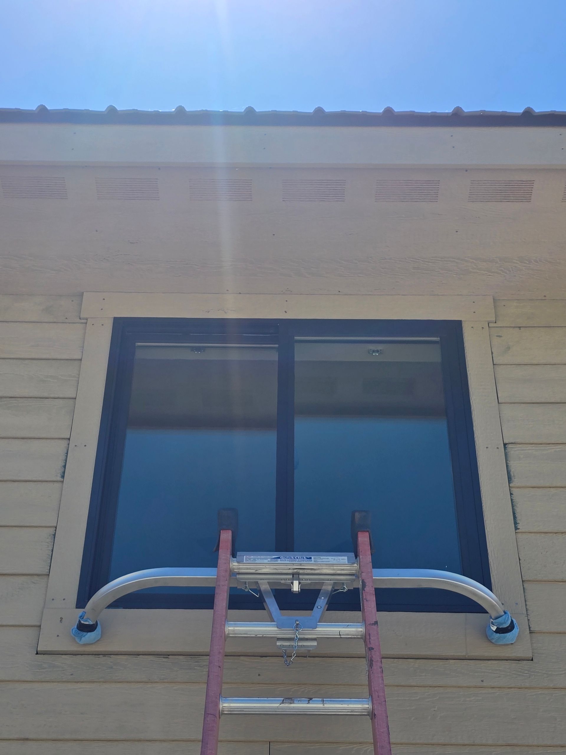 A ladder stabilizer bar is attached to the top of an extension ladder positioned against a residential window.