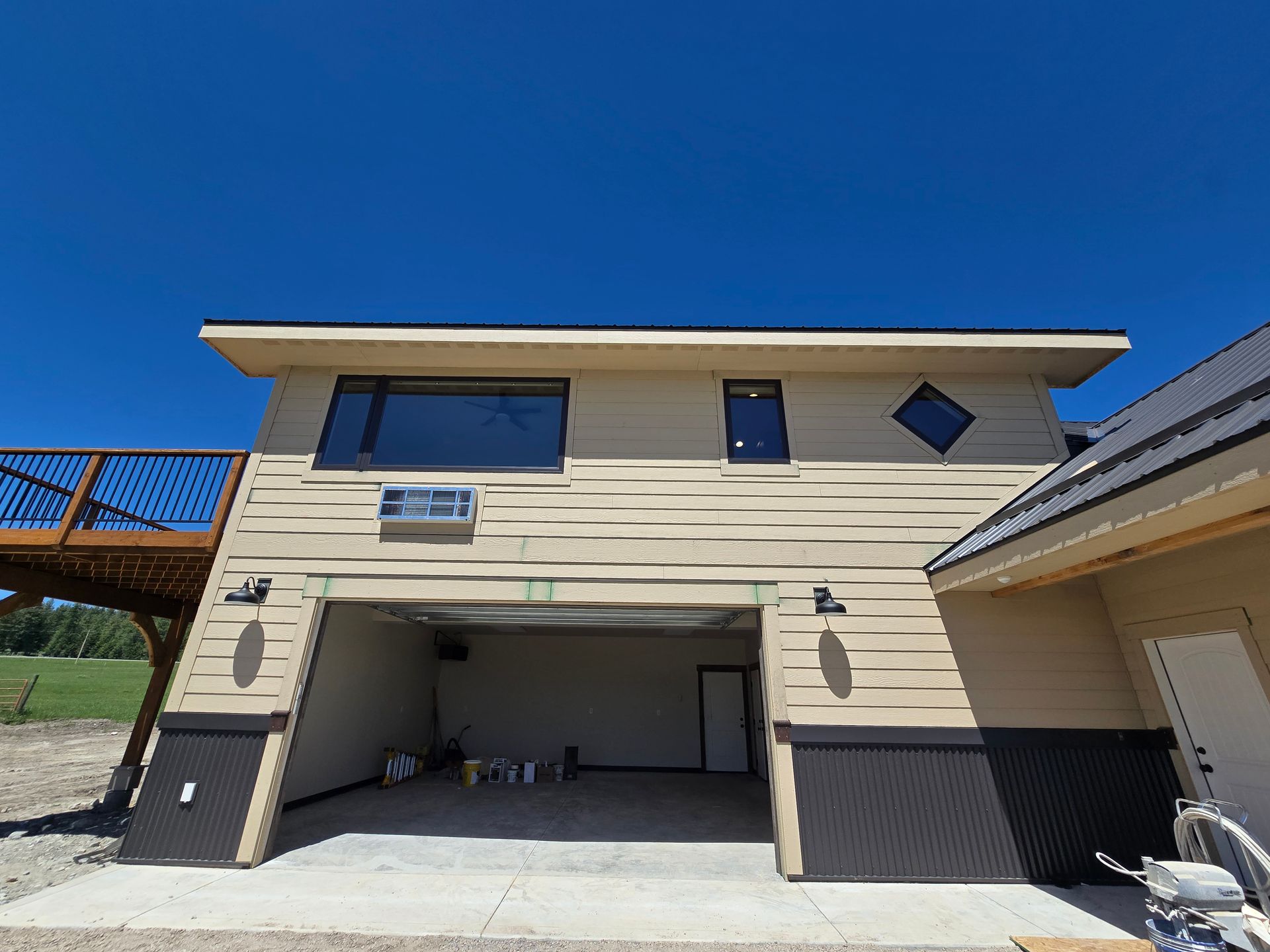 Tan-sided building with a garage bay, upper windows, a wooden deck on the left, and a white door on the right.