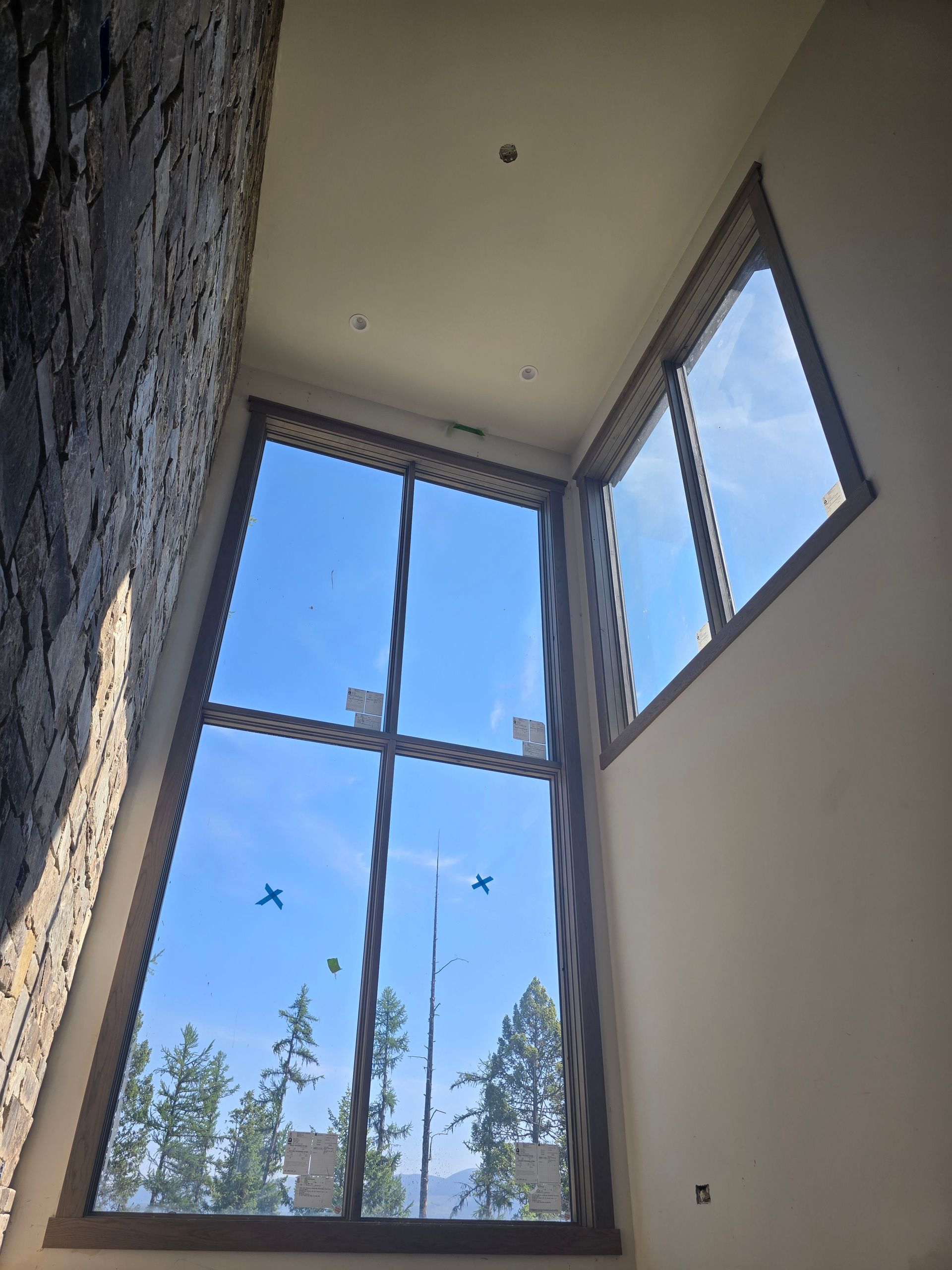 A low-angle view of two large, modern windows set in a white wall next to a textured stone wall, framing a blue sky.