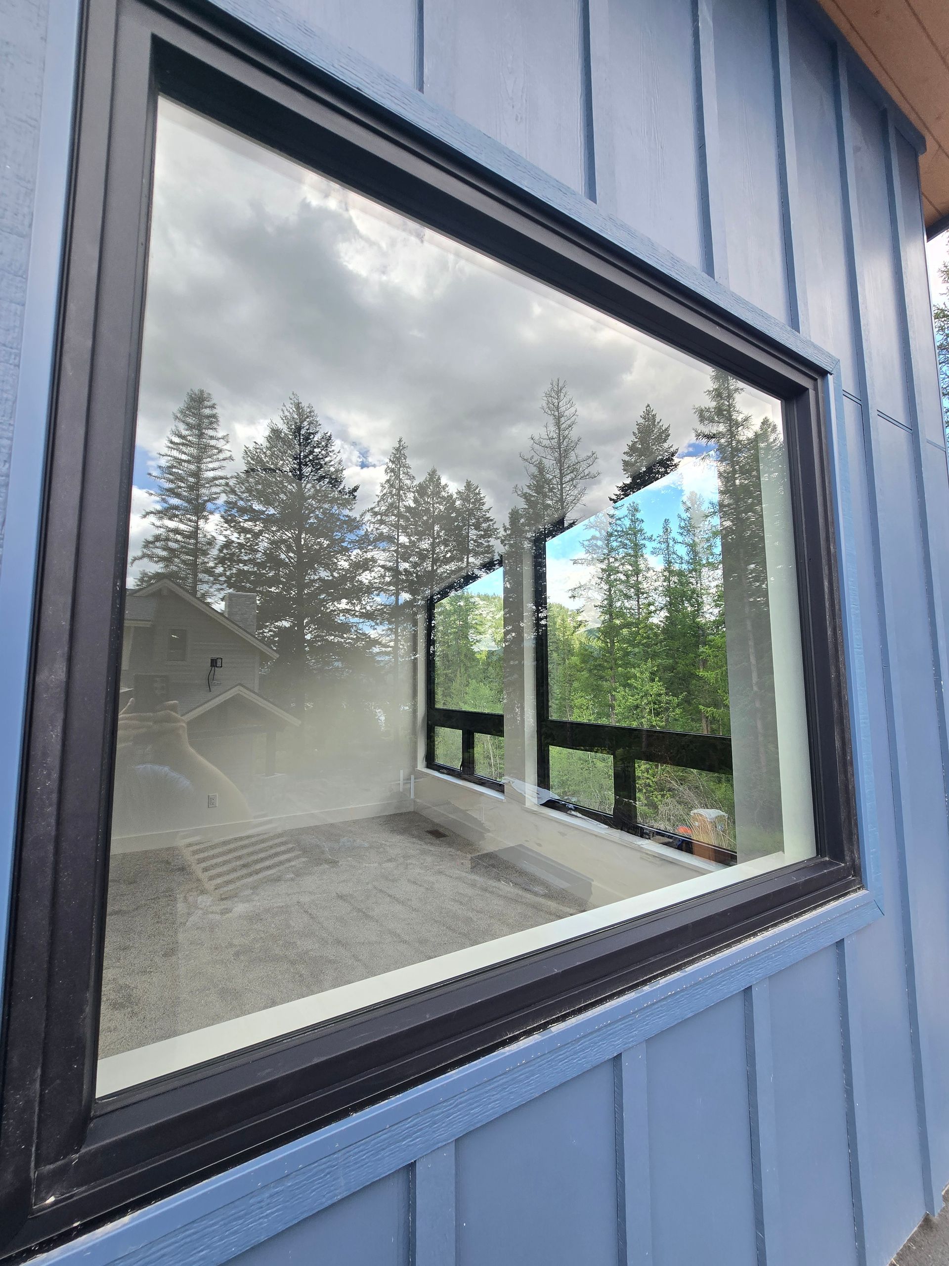 A large square window with a black frame on blue vertical siding, reflecting trees and sky.