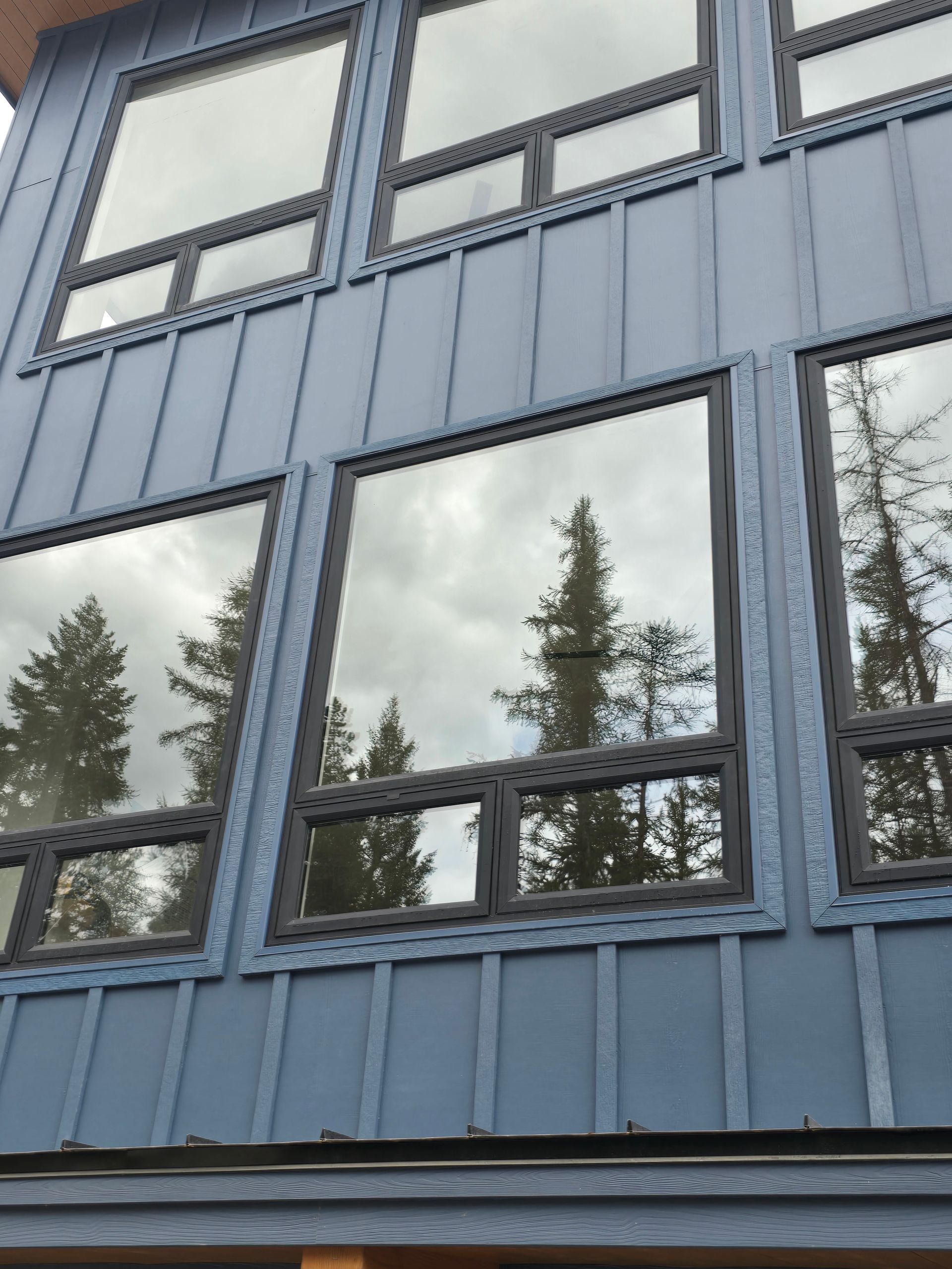 Modern house exterior with blue vertical siding and large windows reflecting pine trees and a cloudy sky.