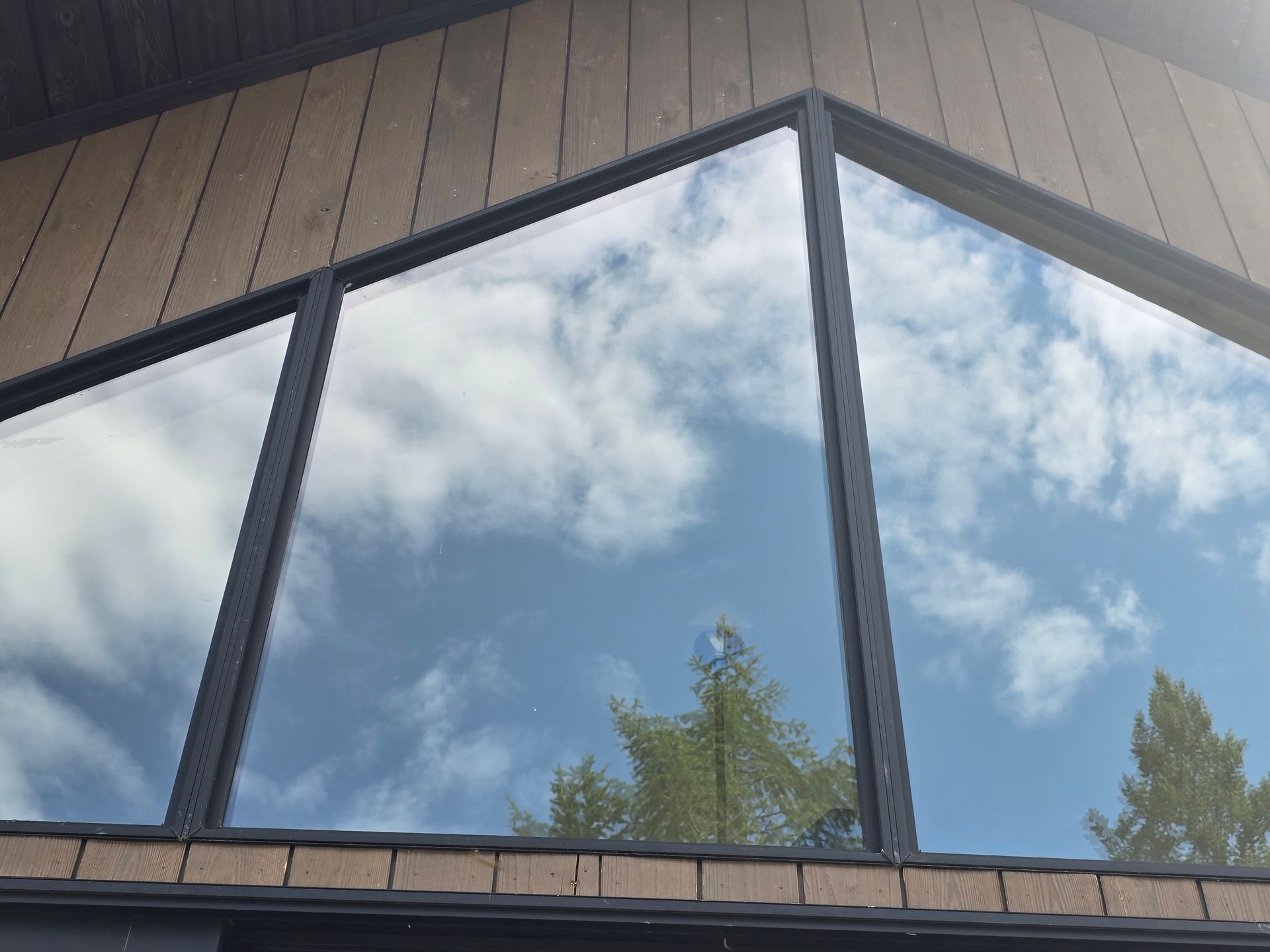 Large triangular window framed in black, reflecting a bright blue sky with clouds and silhouettes of evergreen trees.